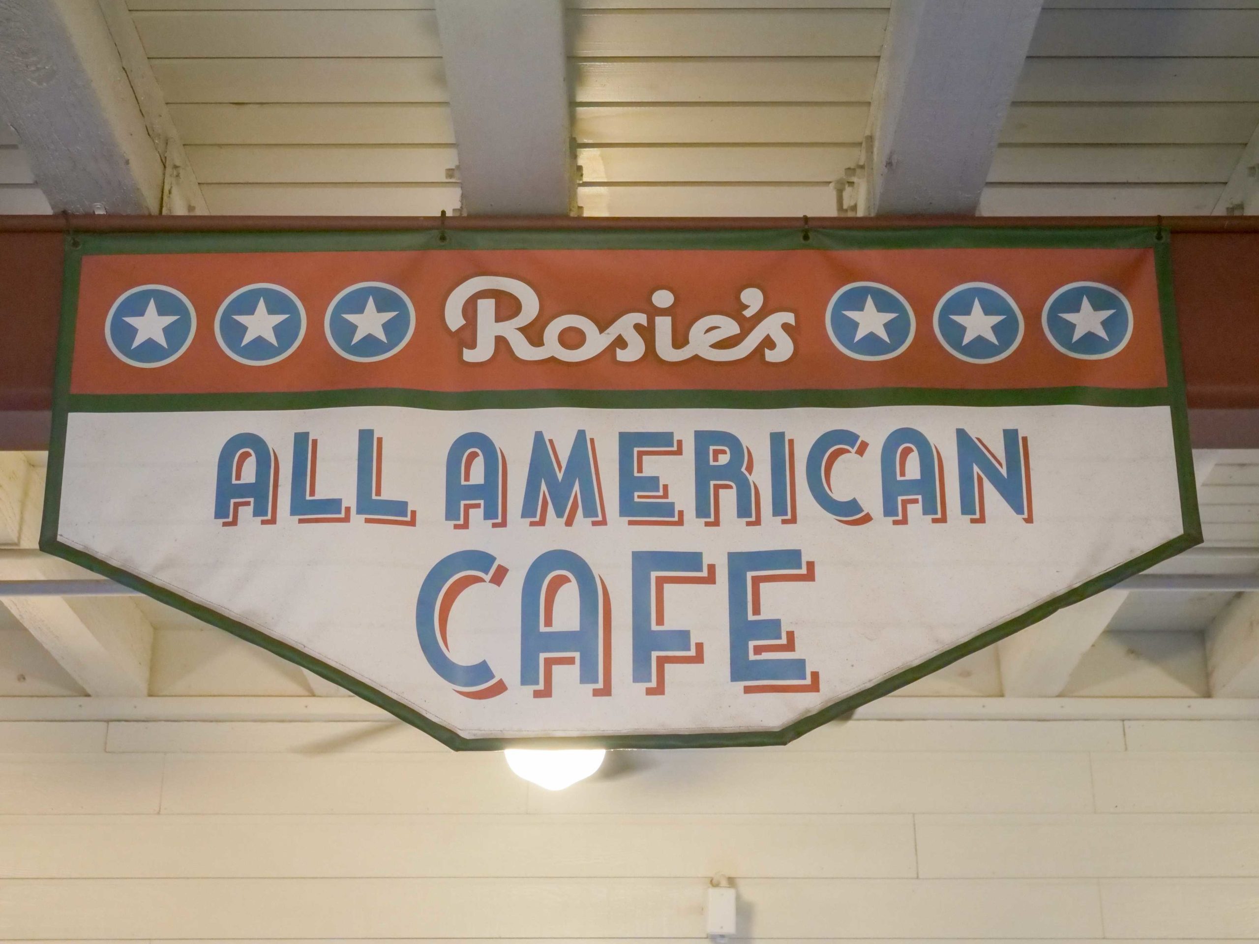 Sign reading "Rosie's All American Cafe" with stars on either side, hanging from a ceiling beam.
