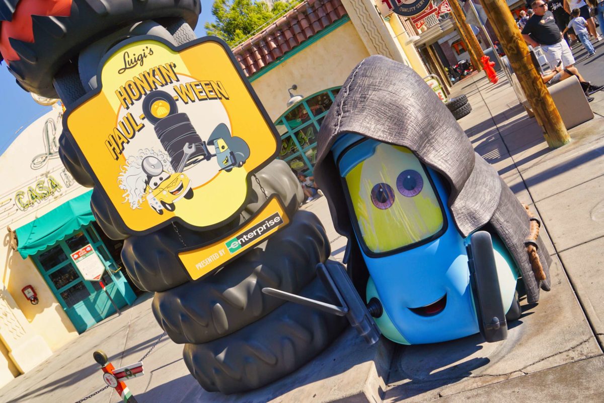 Guido, a blue pitty vehicle with eyes, wears a black cloak. He's in front of a tire stack with a "Luigi's Honkin' Haul-o-Ween" sign.