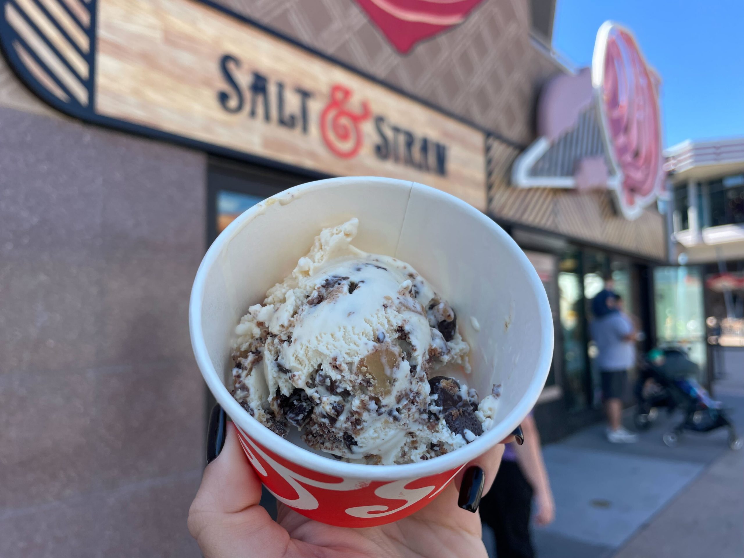 Hand holding a Salt & Straw ice cream cup with a scoop of The Great Candycopia salted butterscotch ice cream with chocolate candy pieces.