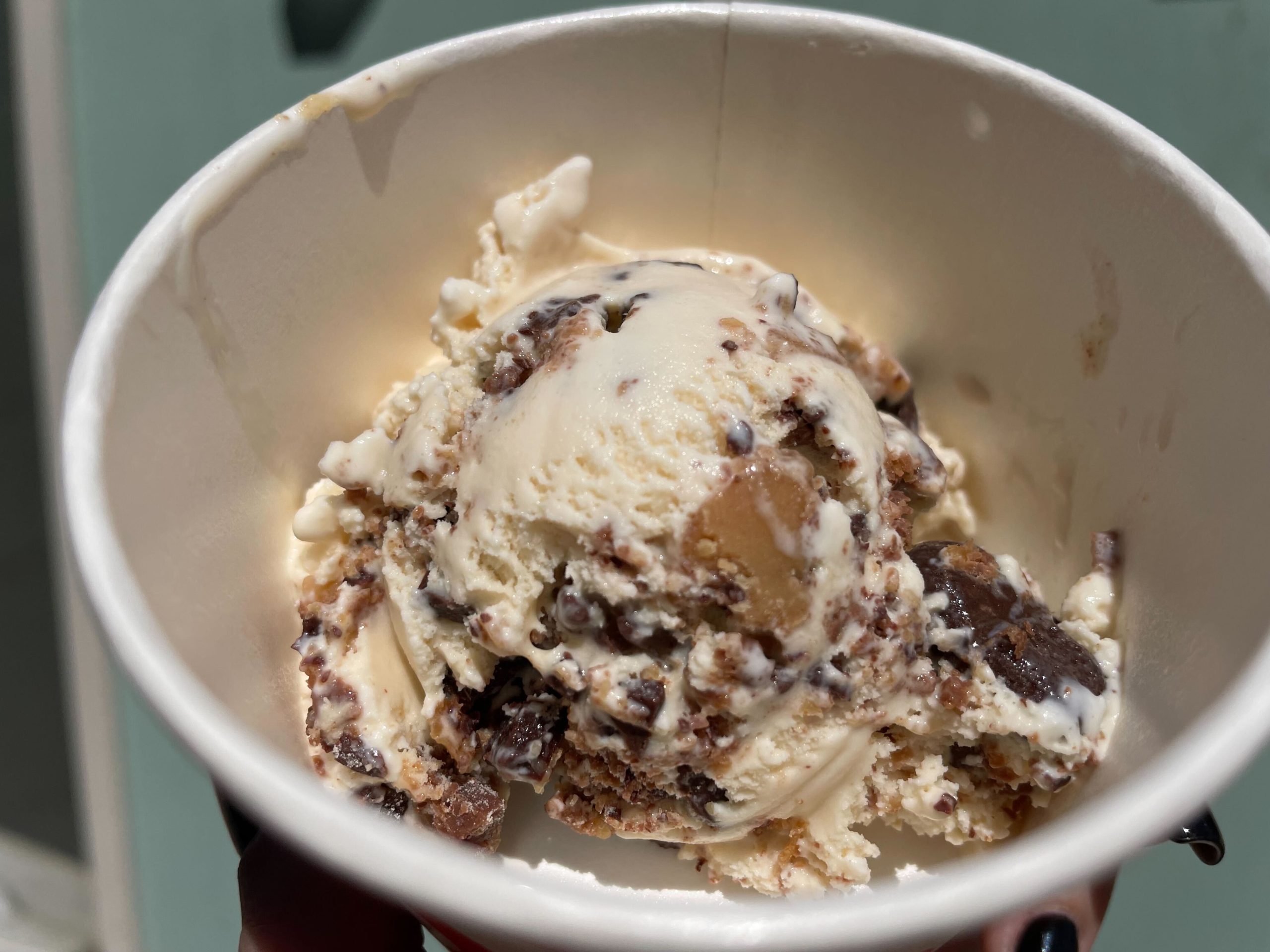 Hand holding a Salt & Straw ice cream cup with a scoop of The Great Candycopia salted butterscotch ice cream with chocolate candy pieces.
