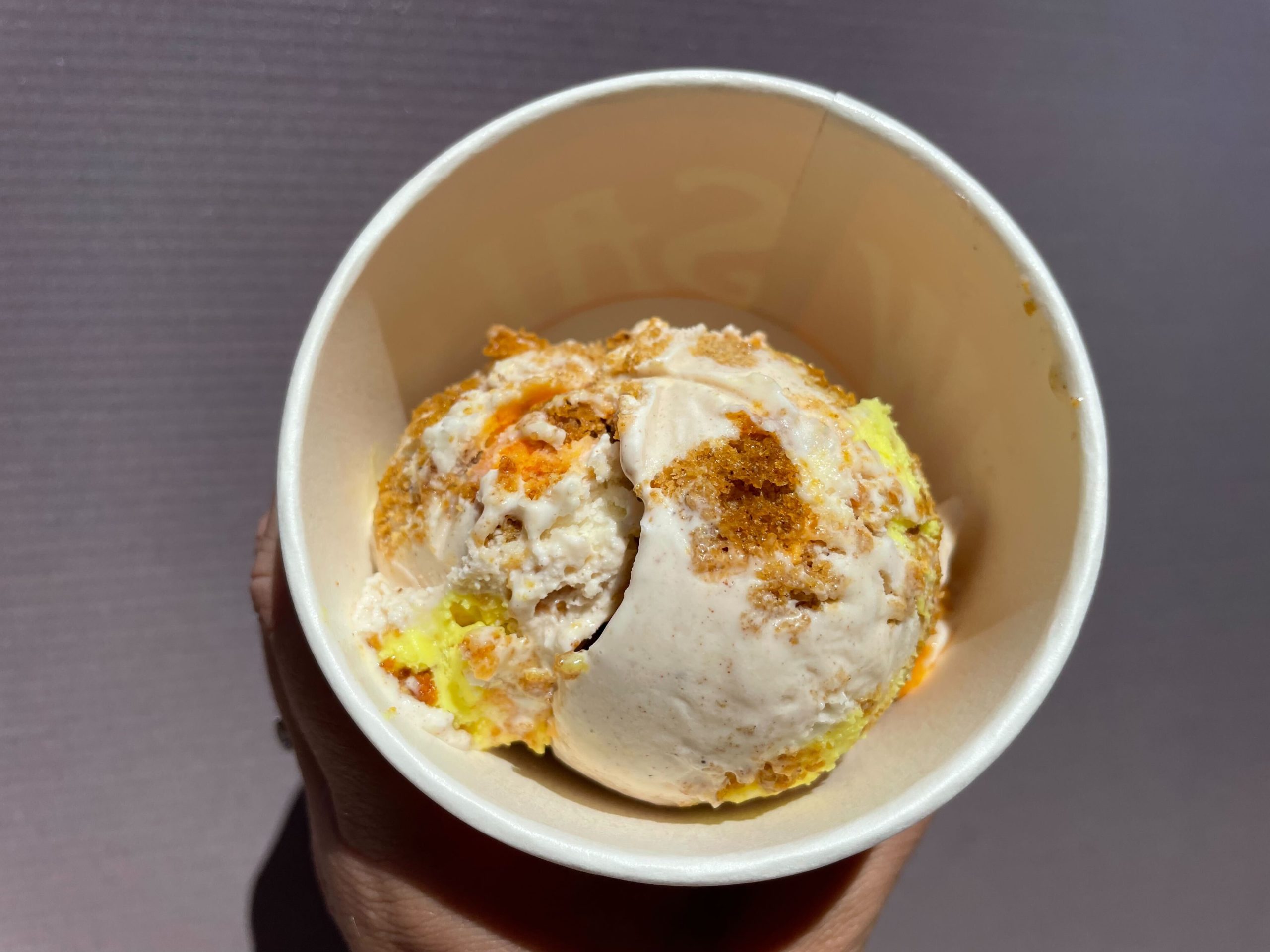 Hand holding a Salt & Straw ice cream cup with a scoop of Jack o' Lantern pumpkin bread ice cream with a cream cheese swirl.