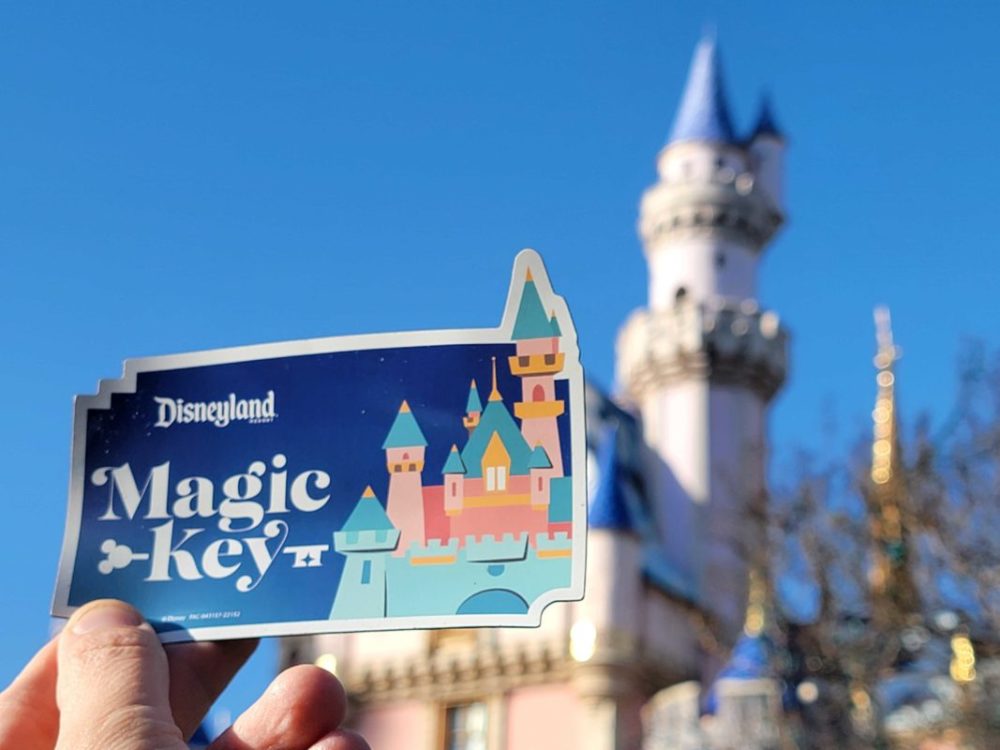 A hand holds a Disneyland Resort Magic Key card in front of a blurred castle against a clear blue sky.