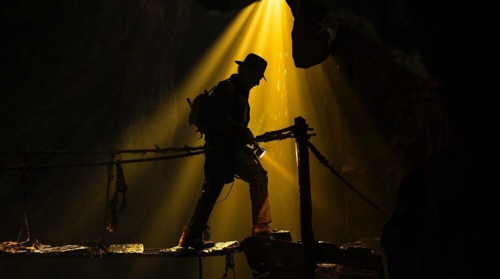 A silhouetted explorer crosses a rope bridge in a dark cave, with a third-quarter beam of light dramatically shining down.