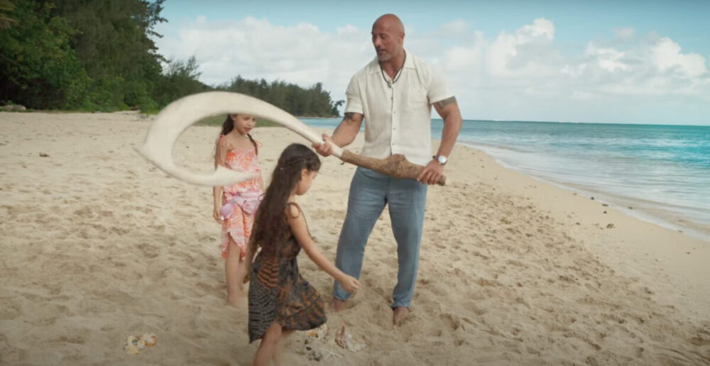 Dwayne Johnson with a real-life Maui fishing hook and his two young daughters, on a beach with the ocean in the background.