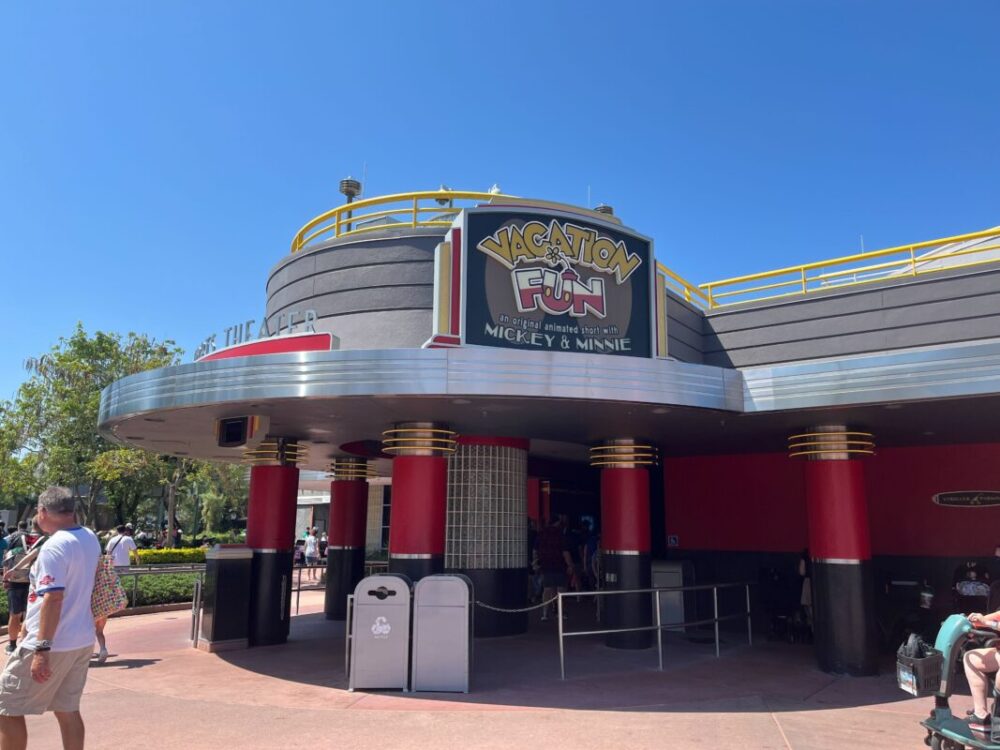 The exterior of a themed attraction building, named "Vacation Fun," featuring Mickey & Minnie. It has a black, gray, and red color scheme with people walking around the area on a sunny day.