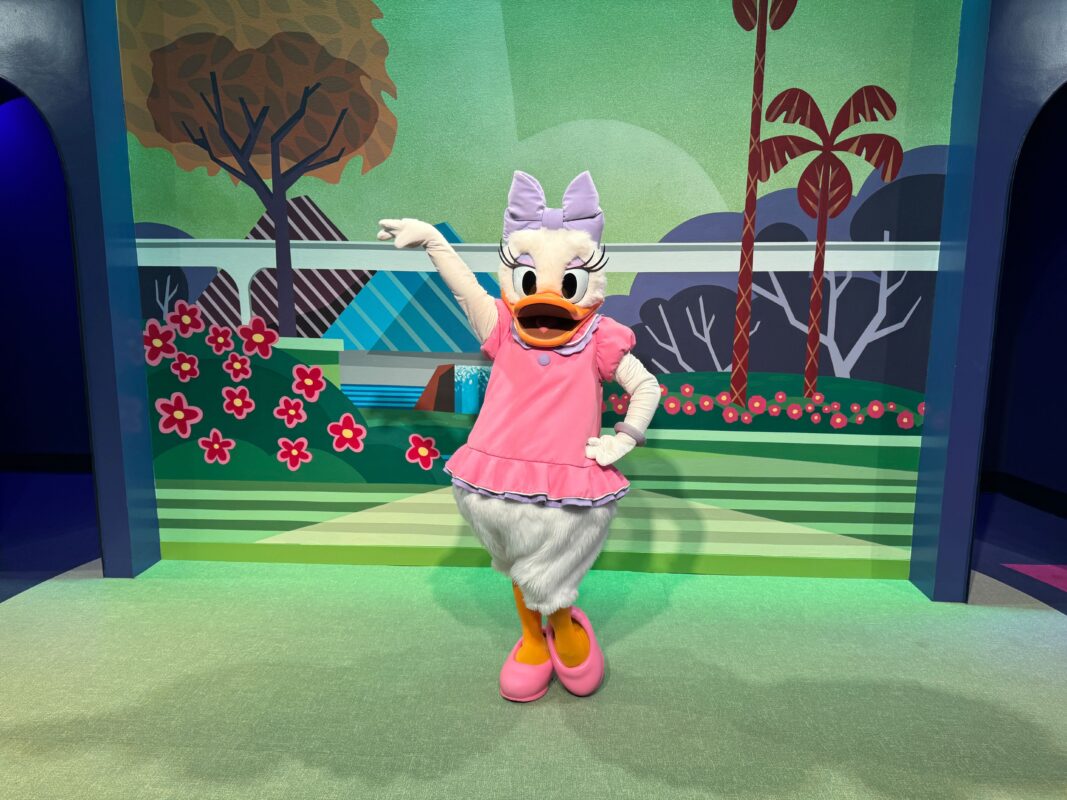 A character in a pink outfit and matching shoes poses with one arm raised in front of a colorful backdrop featuring trees, flowers, and geometric shapes, reminiscent of the vibrant aesthetics often seen at CommuniCore Hall.