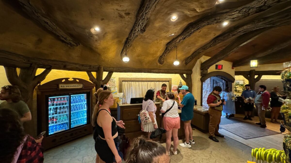 Several people standing and walking inside a rustic-themed room with a wooden ceiling and light fixtures. A refrigerator labeled "Refreshments" is on the left. Near the counter, some folks are interacting, reminiscent of the lively charm of Tiana's Bayou Adventure.