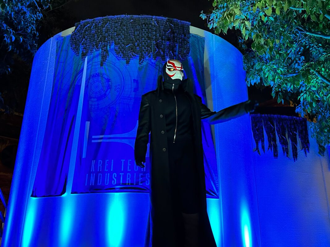 Person in a long, dark coat and mask with red and white patterns standing in front of a lit-up display labeled "Krei Tech Industries." The scene is illuminated with blue lighting and surrounded by foliage.