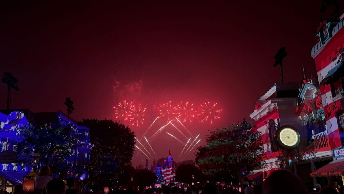 A nighttime fireworks display lights up the sky above a lit castle, with buildings and trees decorated in red, white, and blue lights, and a large clock visible on the right side.