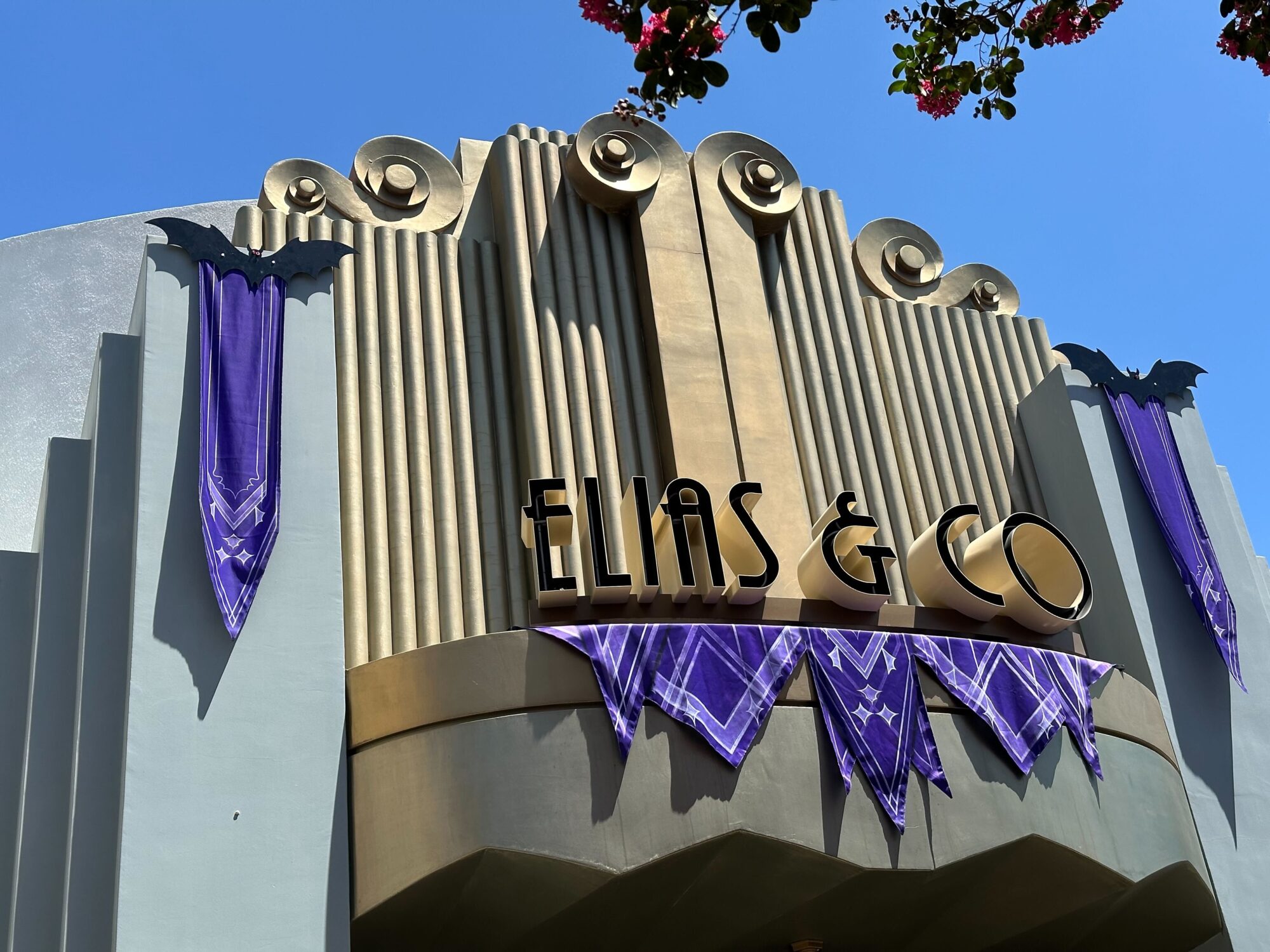 The Art Deco facade of the Elias & Co building at Disneyland Resort is adorned with festive Halloween decor, featuring purple bat-themed decorations against a clear blue sky.
