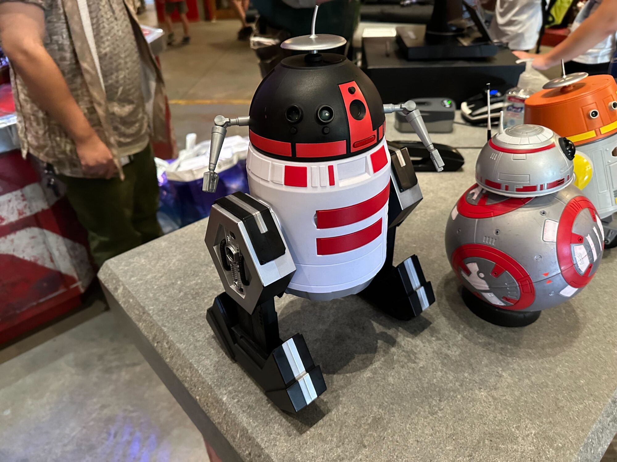 Three robot toy figures are displayed on a gray table, with people standing in the background. One is black and white with red markings, another is silver and red, and a third is partially visible and orange.