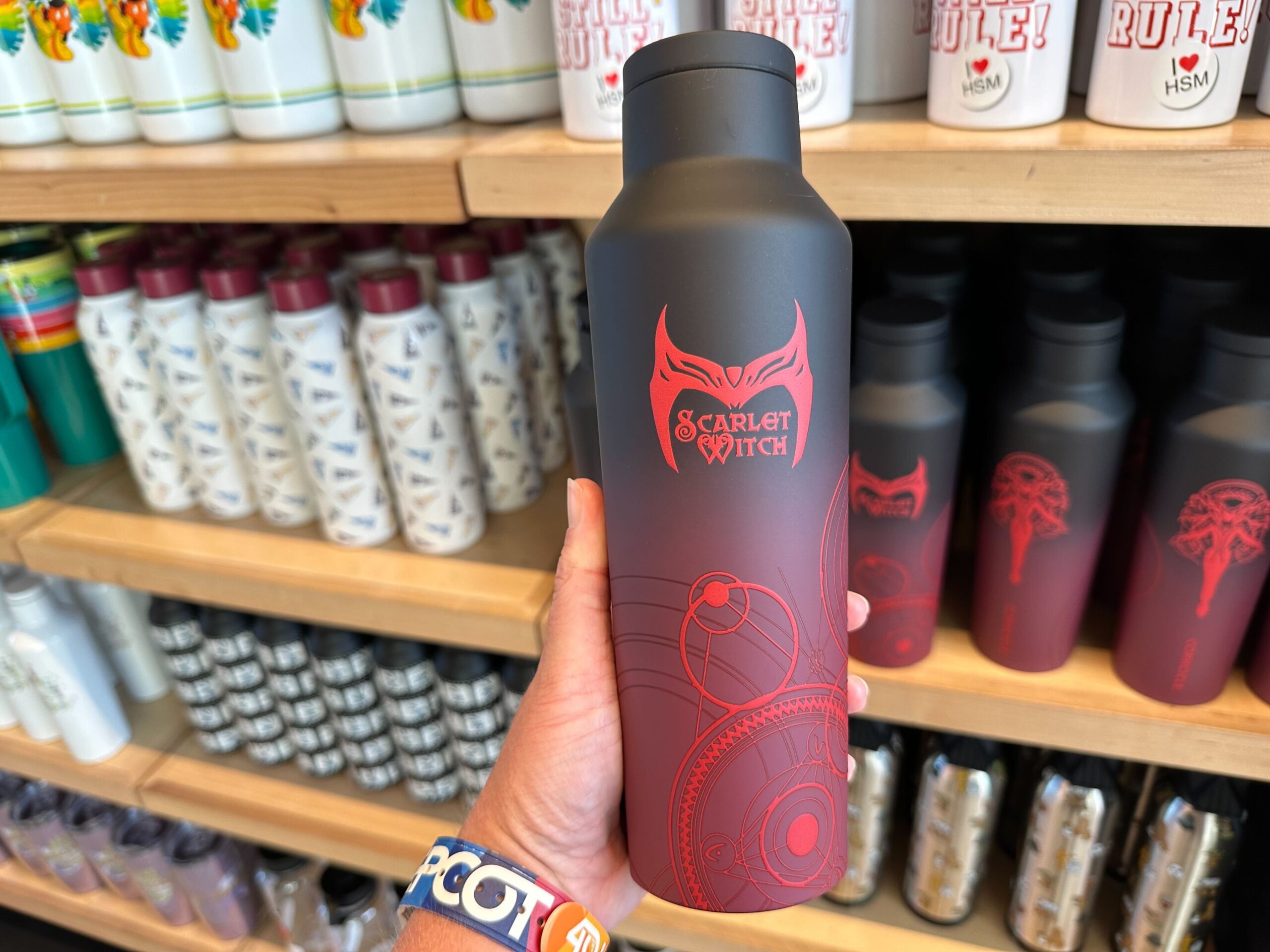 A hand holds a water bottle adorned with "Scarlet Witch" text and graphic, in front of shelves showcasing various other water bottles.