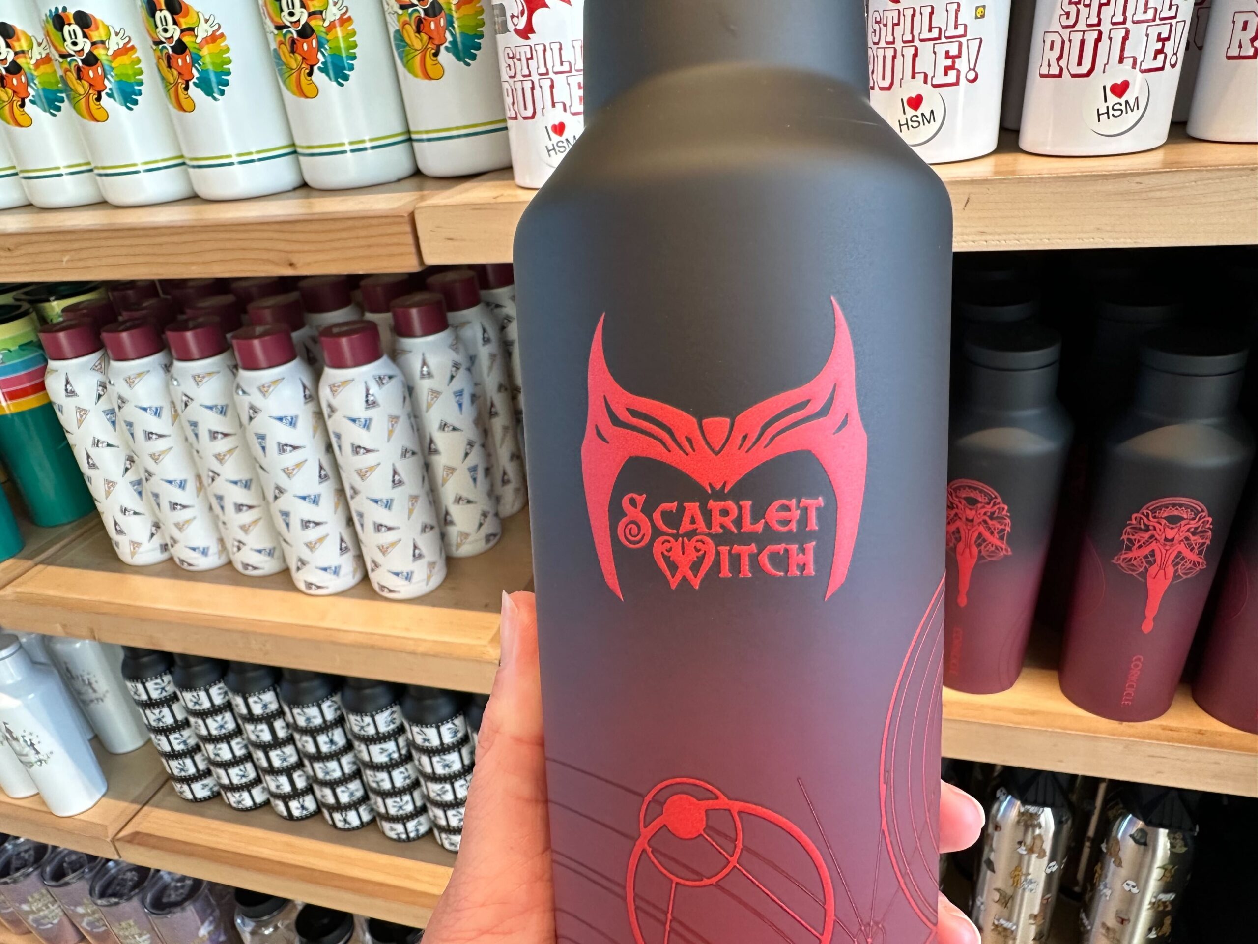 A gray water bottle with a prominent red Scarlet Witch logo is displayed, held in front of a shelf filled with other themed bottles, including ones featuring Mickey Mouse and High School Musical designs.