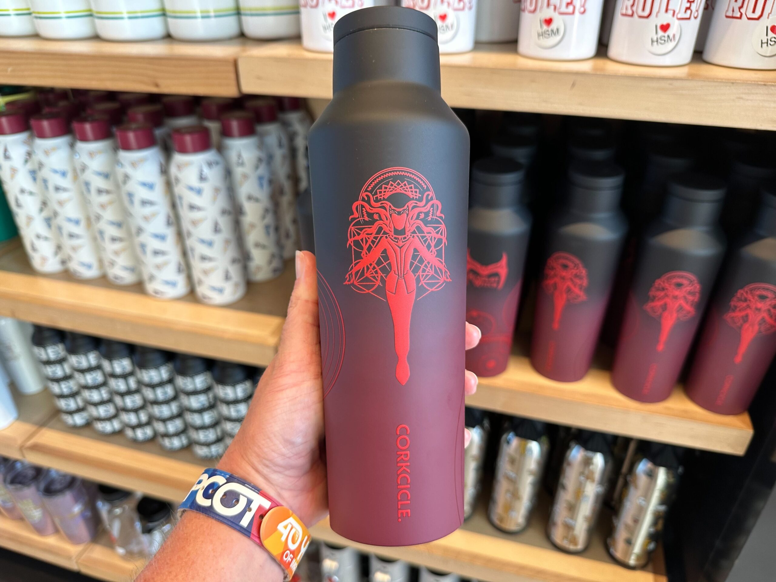 A hand holding a black and red Corkcicle water bottle featuring a Scarlet Witch-inspired ornate design, positioned in front of shelves stocked with similar and assorted water bottles.