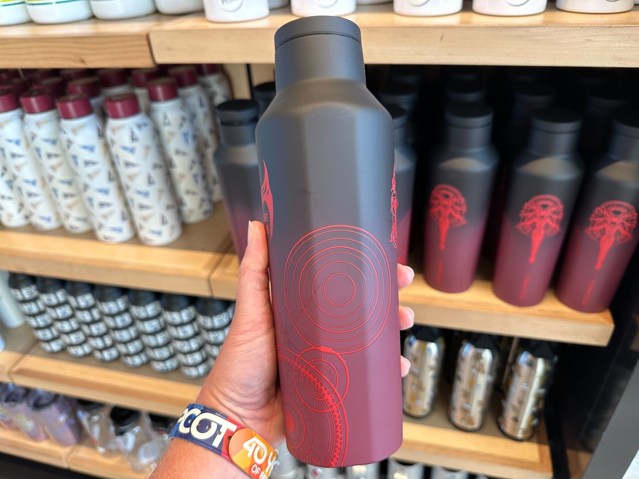 A hand holds a black and red insulated water bottle with circular designs, reminiscent of Scarlet Witch's iconic colors, in front of a store display of various water bottles.