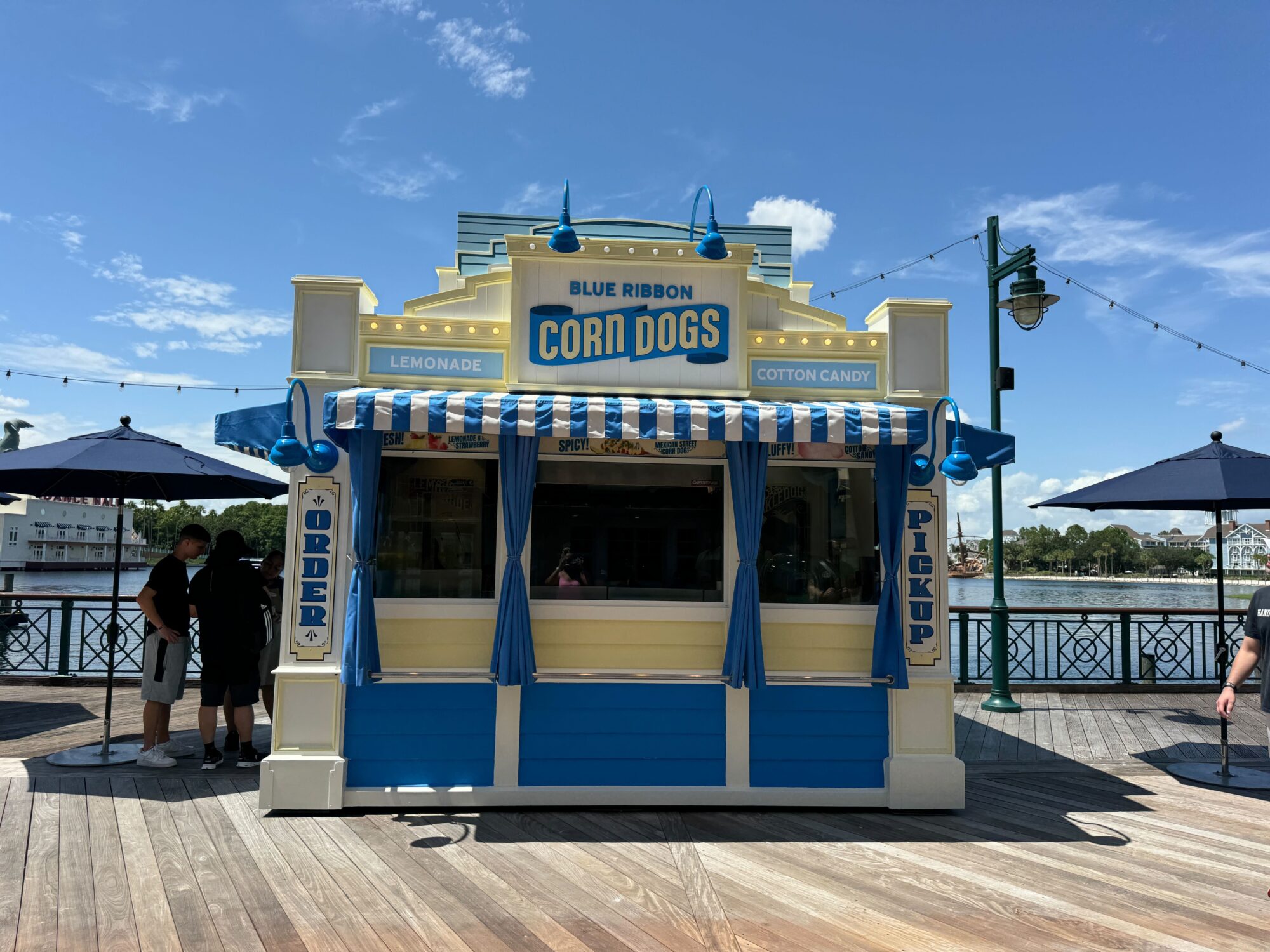 A blue and white food stand named "Blue Ribbon Corn Dogs" offers lemonade and cotton candy. There are two labeled windows for ordering and pickup, and it's situated by a waterfront under a clear sky.