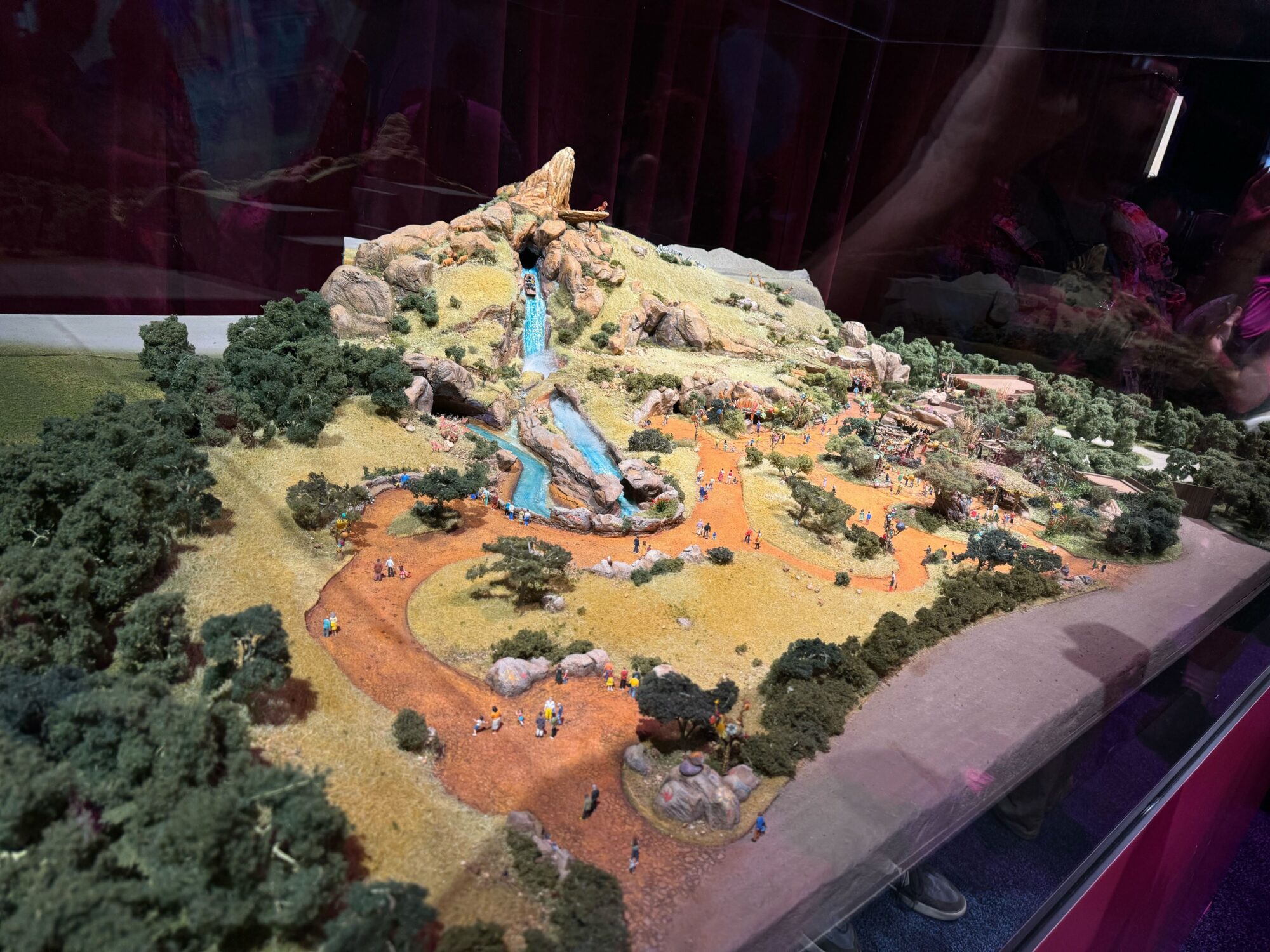 A detailed scale model of a theme park attraction featuring a mountain with cascading water, winding pathways, trees, and tiny figurines of people. Inspired by The Lion King, the display captures the magic and adventure of the savannah in a glass enclosure.