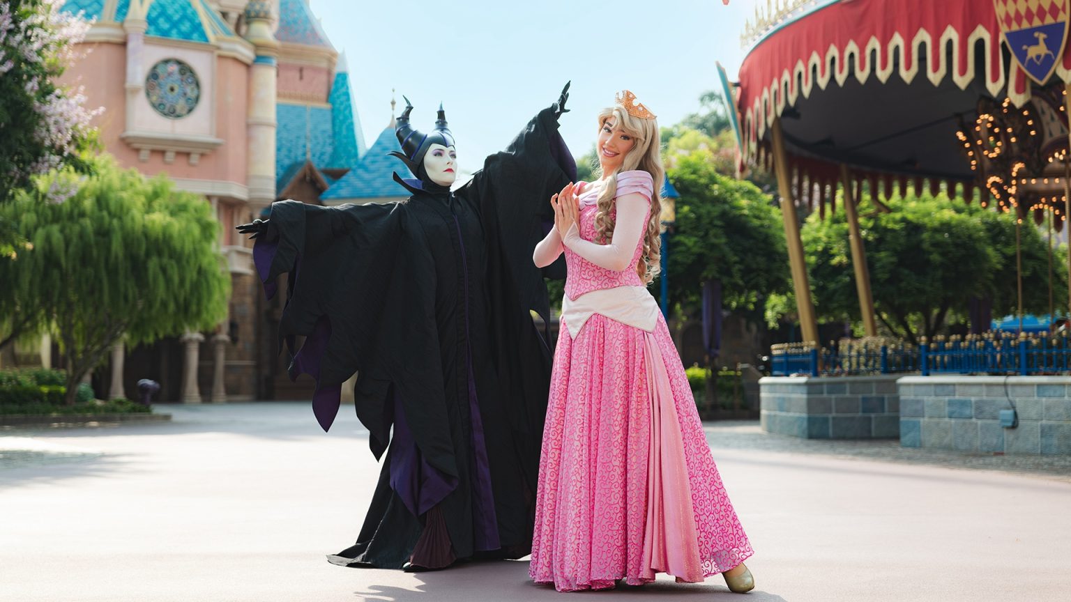 Two people in Disney character costumes: Maleficent in black and Aurora in a pink dress, pose in an outdoor theme park setting with rides and a castle in the background.