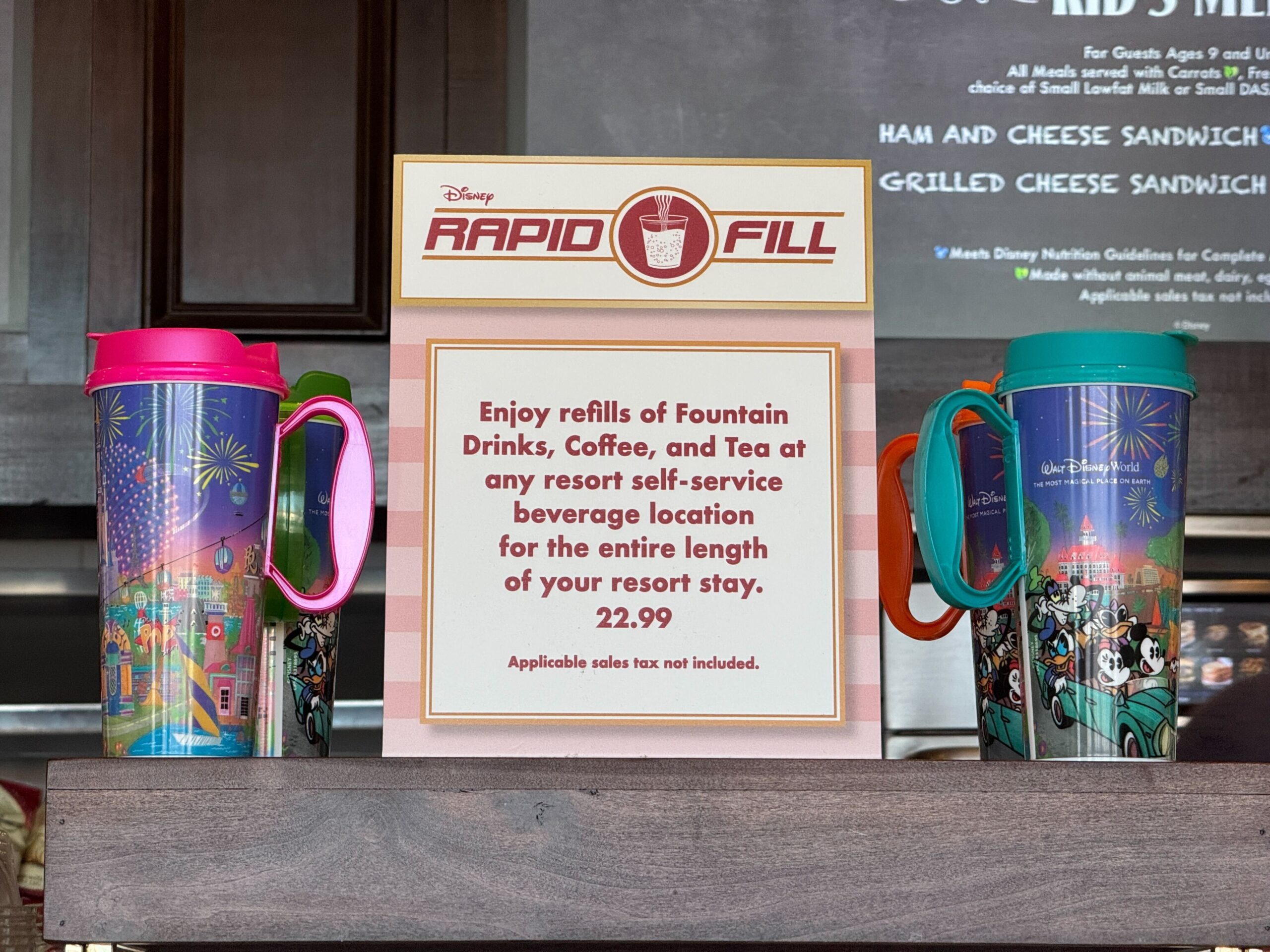 Two colorful Resort Refillable Mugs are displayed beside a sign at Walt Disney World. The sign offers unlimited refills of drinks at resort locations for the length of stay, priced at $22.99. Take advantage before prices increase!