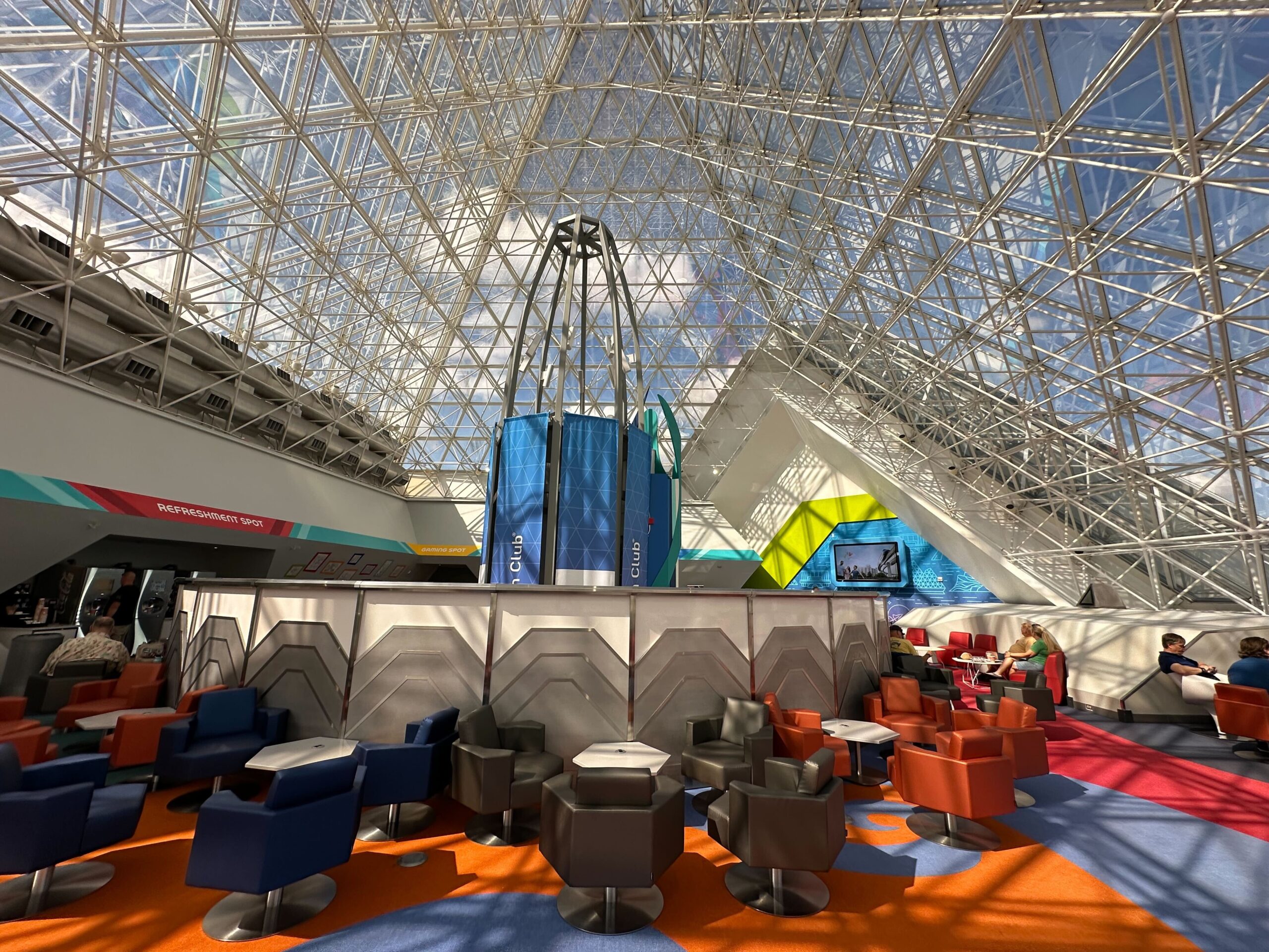 Modern lounge with colorful seating under a geometric glass ceiling, featuring a central structure with screens displaying blue graphics. Bright sunlight streams through the roof.
