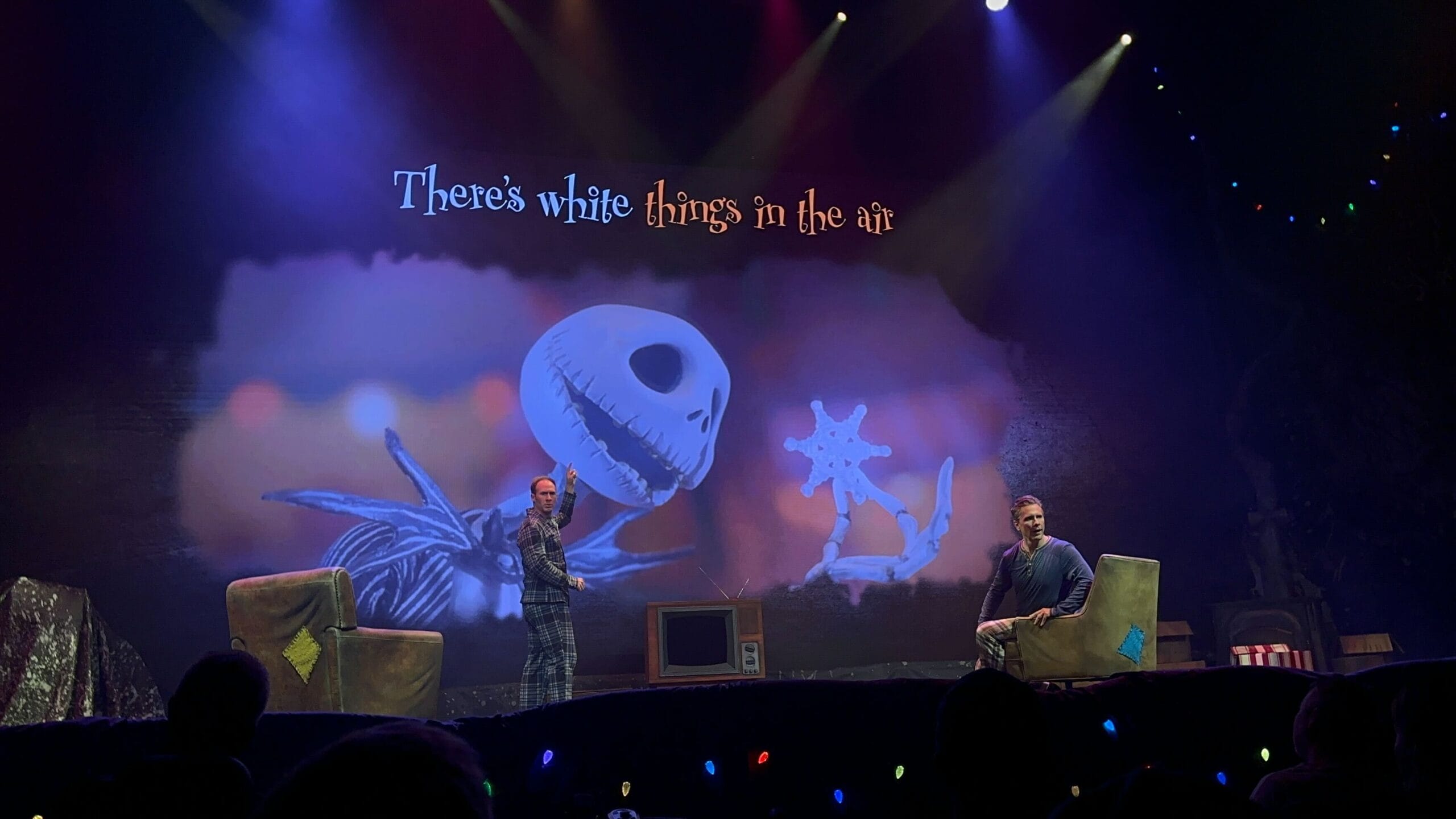 Two people sit on stage with a colorful backdrop featuring a skeleton character and the words "There’s white things in the air." The Nightmare Before Christmas Sing-Along comes to life as stage lighting adds a dramatic effect.