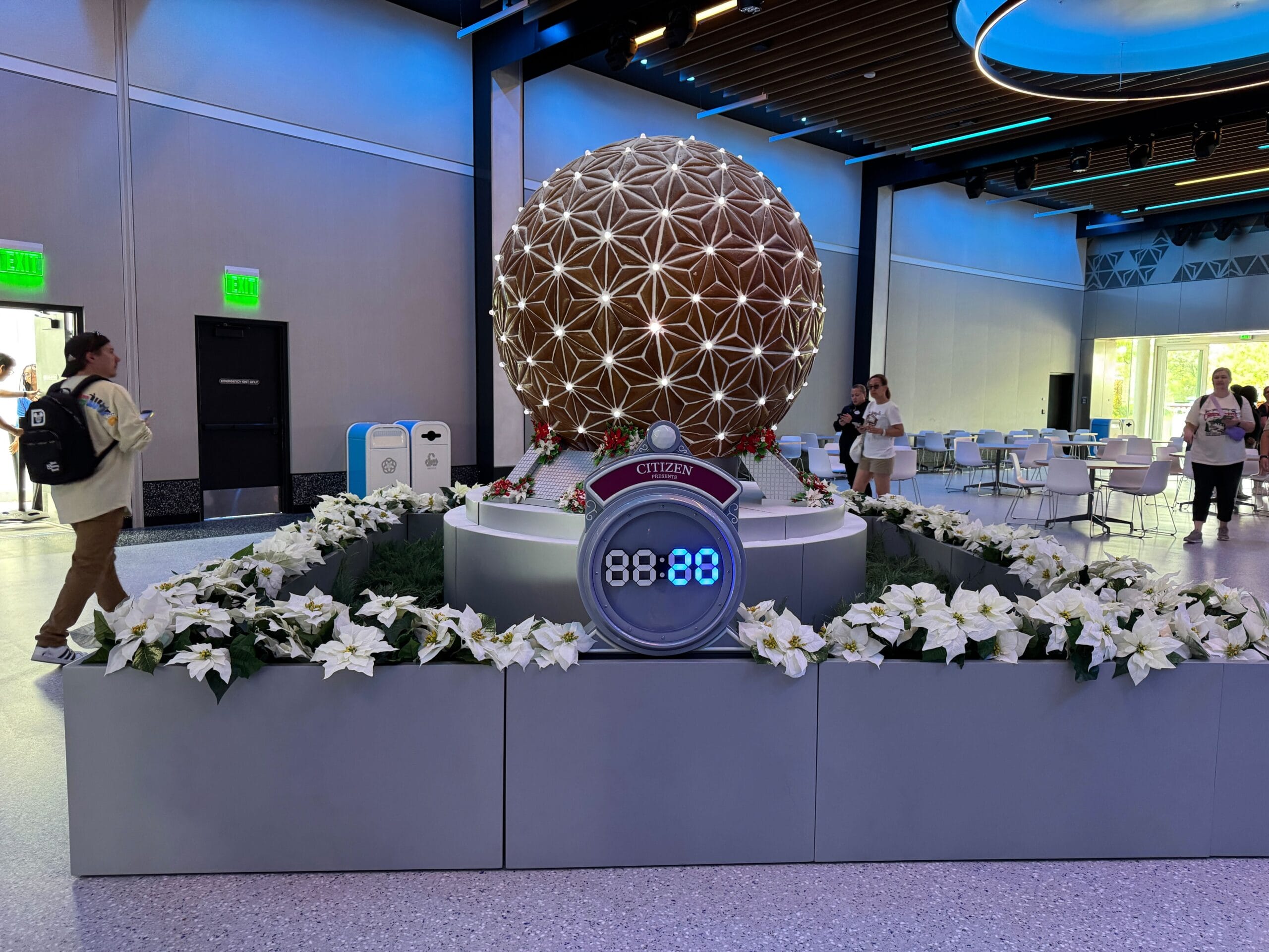 A decorative globe resembling a gingerbread spaceship, along with a digital clock displaying "88:88," is surrounded by a white flower arrangement in a modern indoor setting. People are walking nearby.