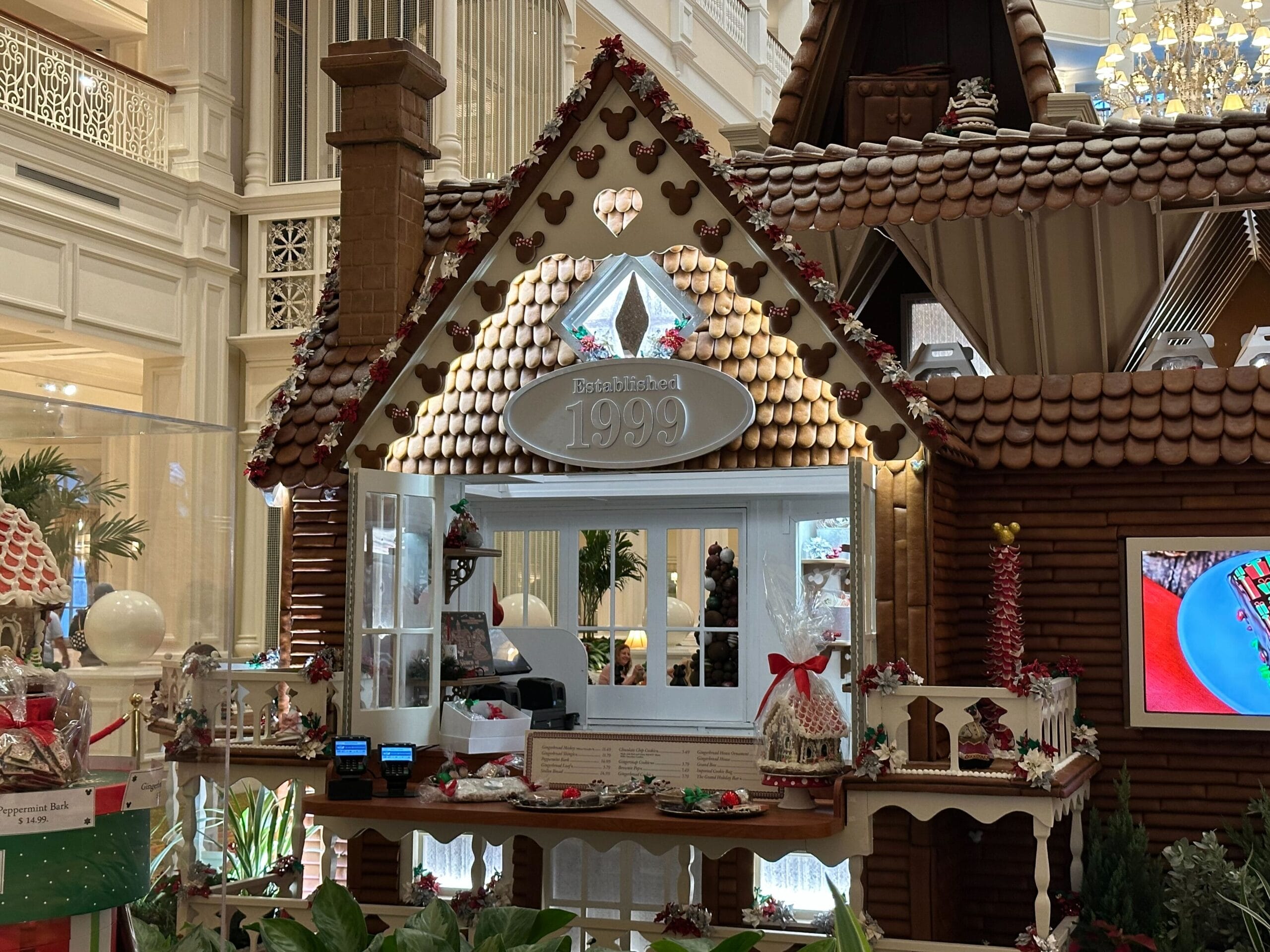 A Grand Floridian Gingerbread House display showcases a sign reading "Established 1999." It features festive decorations, a counter with wrapped treats, and a TV screen displaying vibrant images.