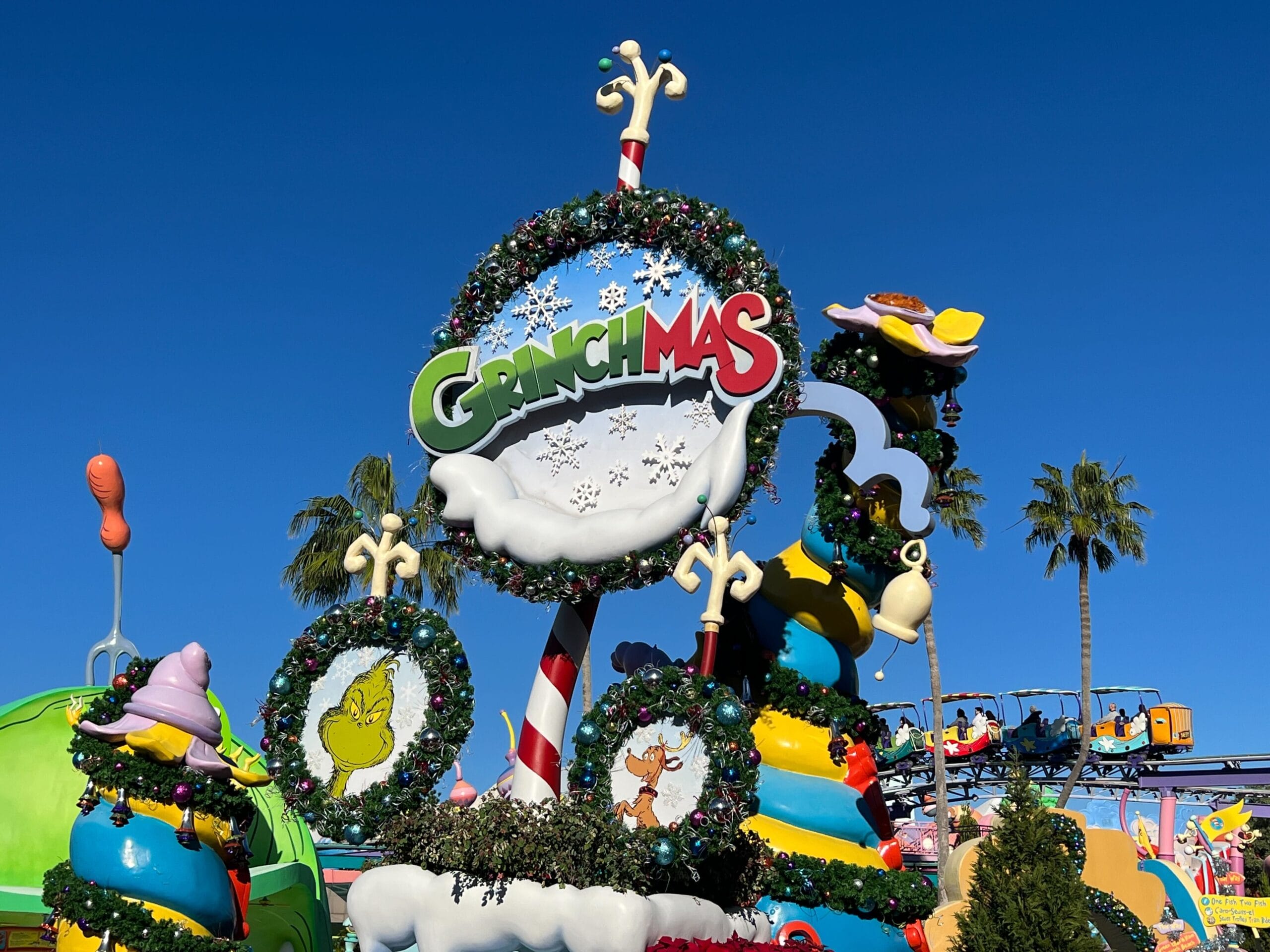 Festive Grinch-themed decorations with Christmas wreaths, a snow-covered sign reading "Grinchmas," and palm trees in the background under a clear blue sky.