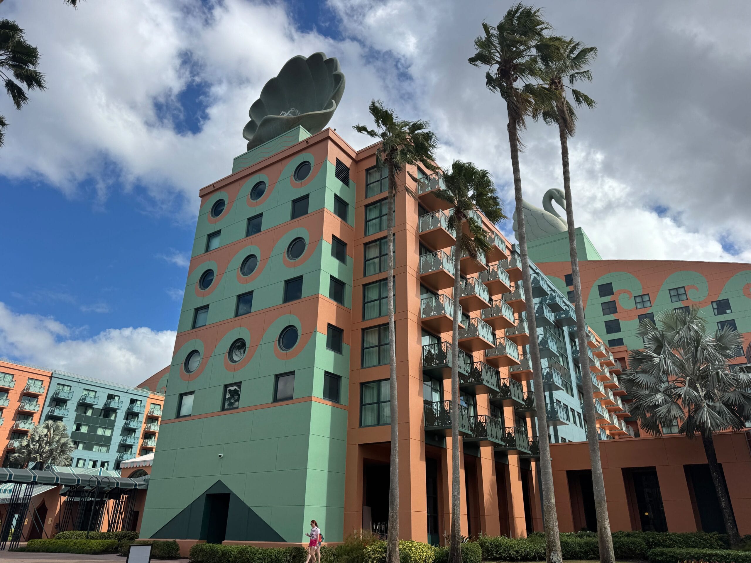 A multi-story building with teal and orange geometric patterns, circular windows, palm trees in the foreground, and a large decorative seashell on the roof.