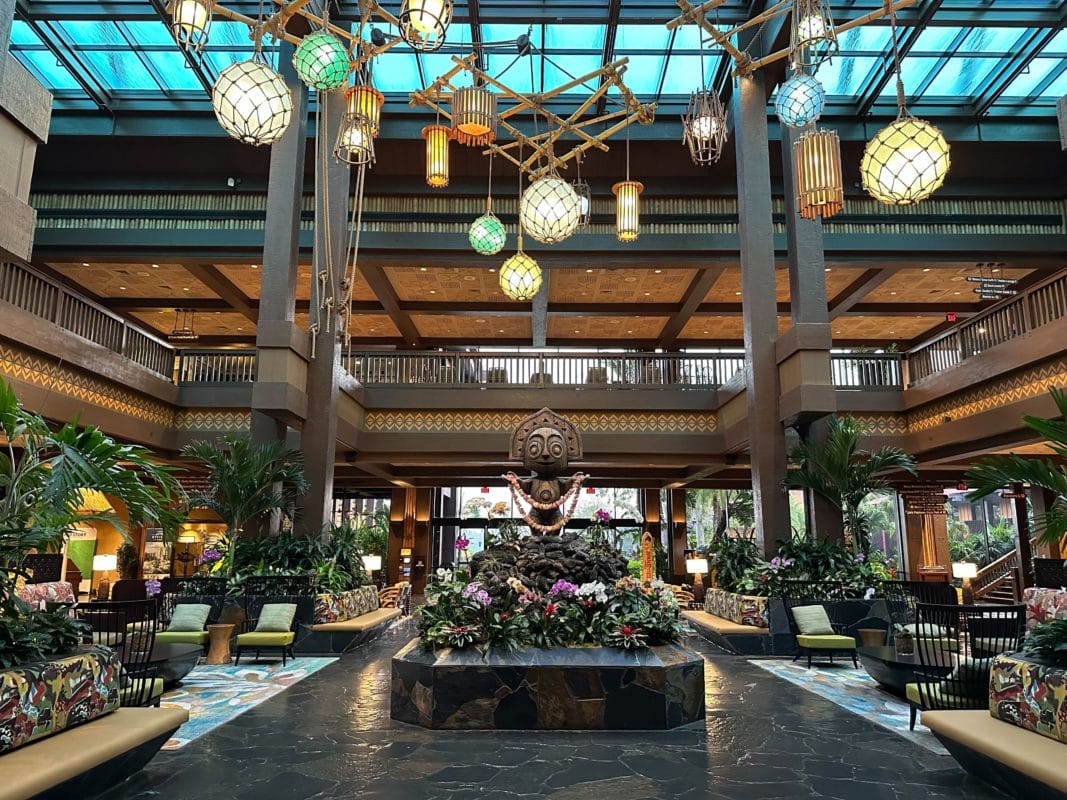 The spacious Polynesian Village Resort lobby features tall columns, a central sculpture, and hanging lights. Floral and plant decorations enhance the charm, with cozy seating areas on either side beneath a sky ceiling with a grid pattern.