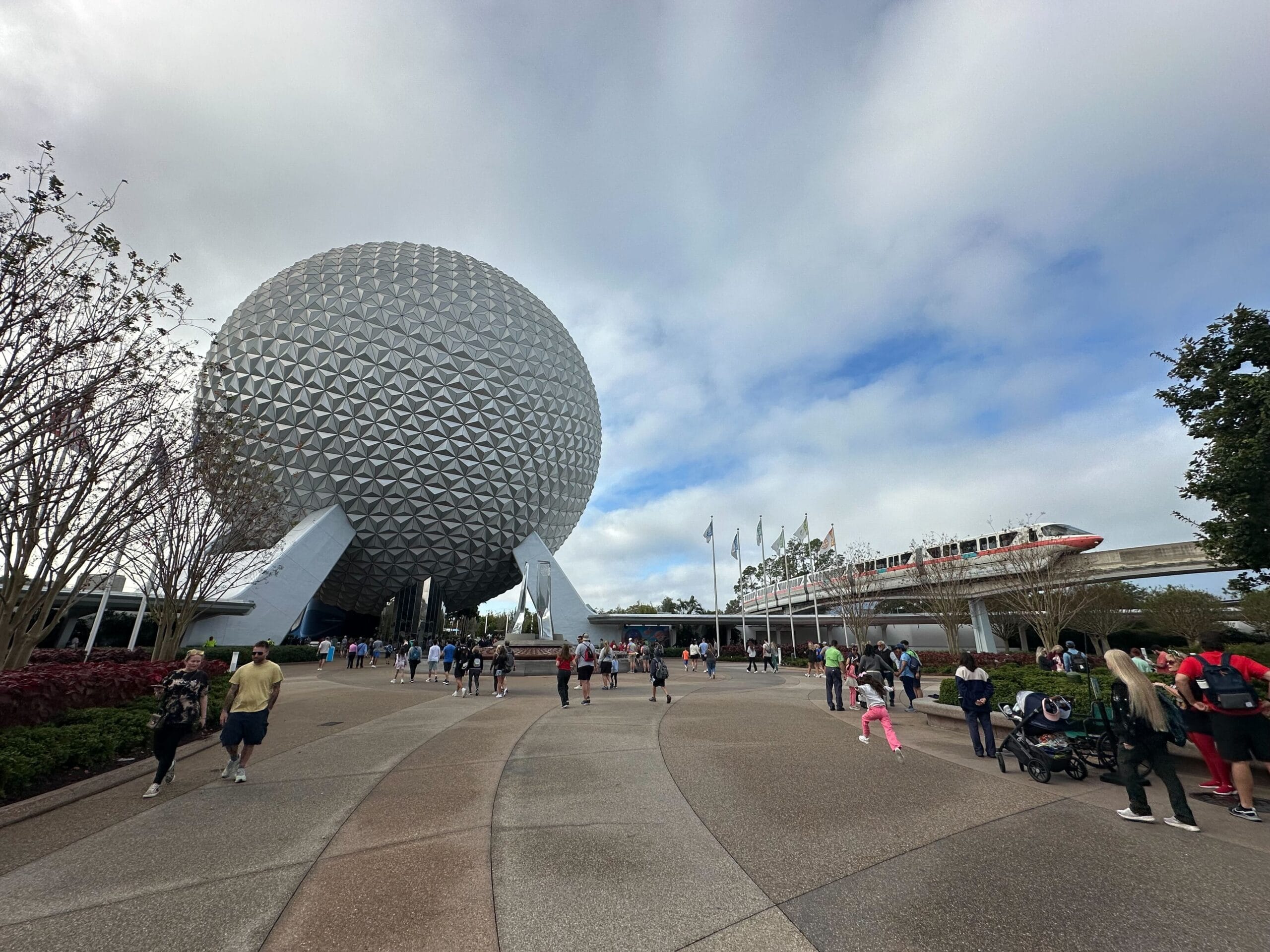 People walk near the large geodesic sphere of Epcot, with a monorail passing by and a cloudy sky above.