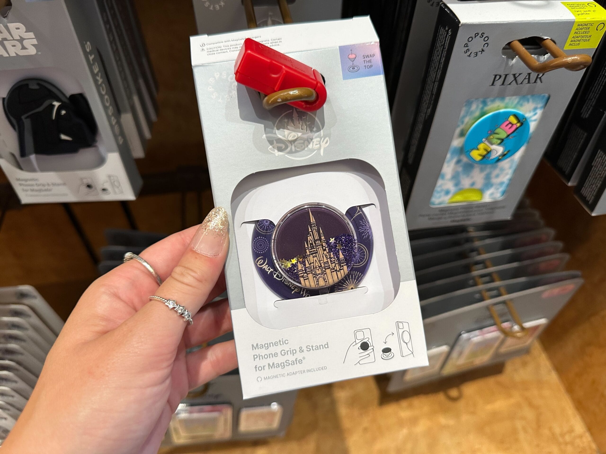 Hand holding a Disney magnetic phone grip with Cinderella Castle design, displayed next to Disney Ears on a store shelf.