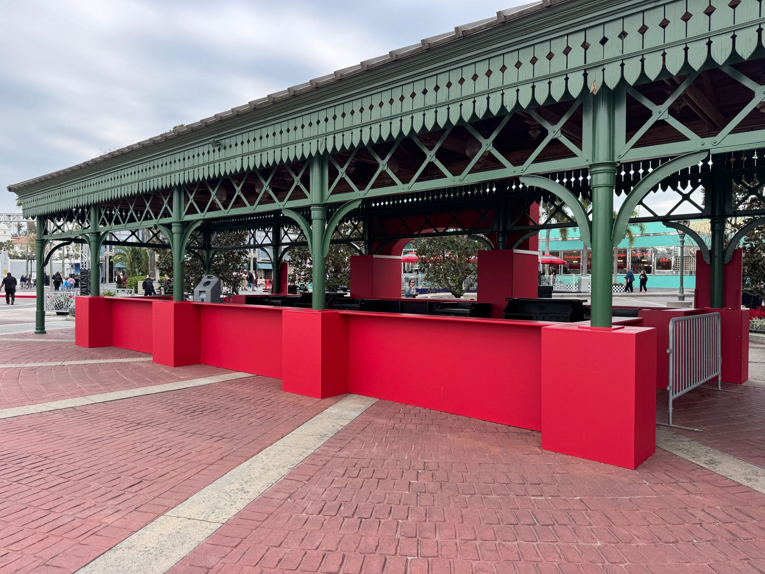 A red and green outdoor pavilion, reminiscent of a Mardi Gras celebration, stands with its covered roof and open counter spaces in a plaza with brick flooring under a cloudy sky. Nearby food booths draw people as they walk in the background.