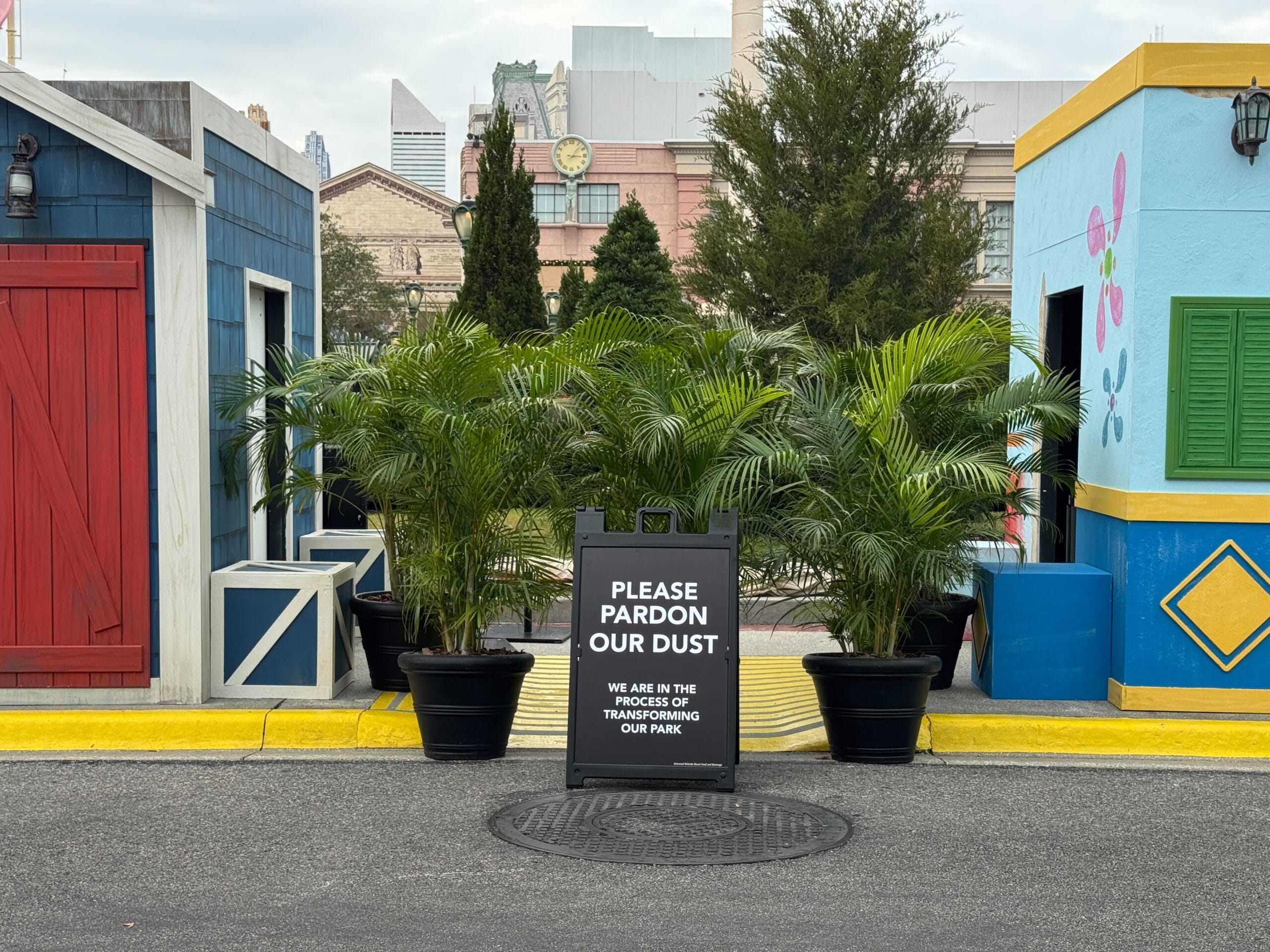 Sign reading "Please pardon our dust. We are in the process of transforming our park for Mardi Gras festivities" placed on a street surrounded by colorful buildings and potted plants.