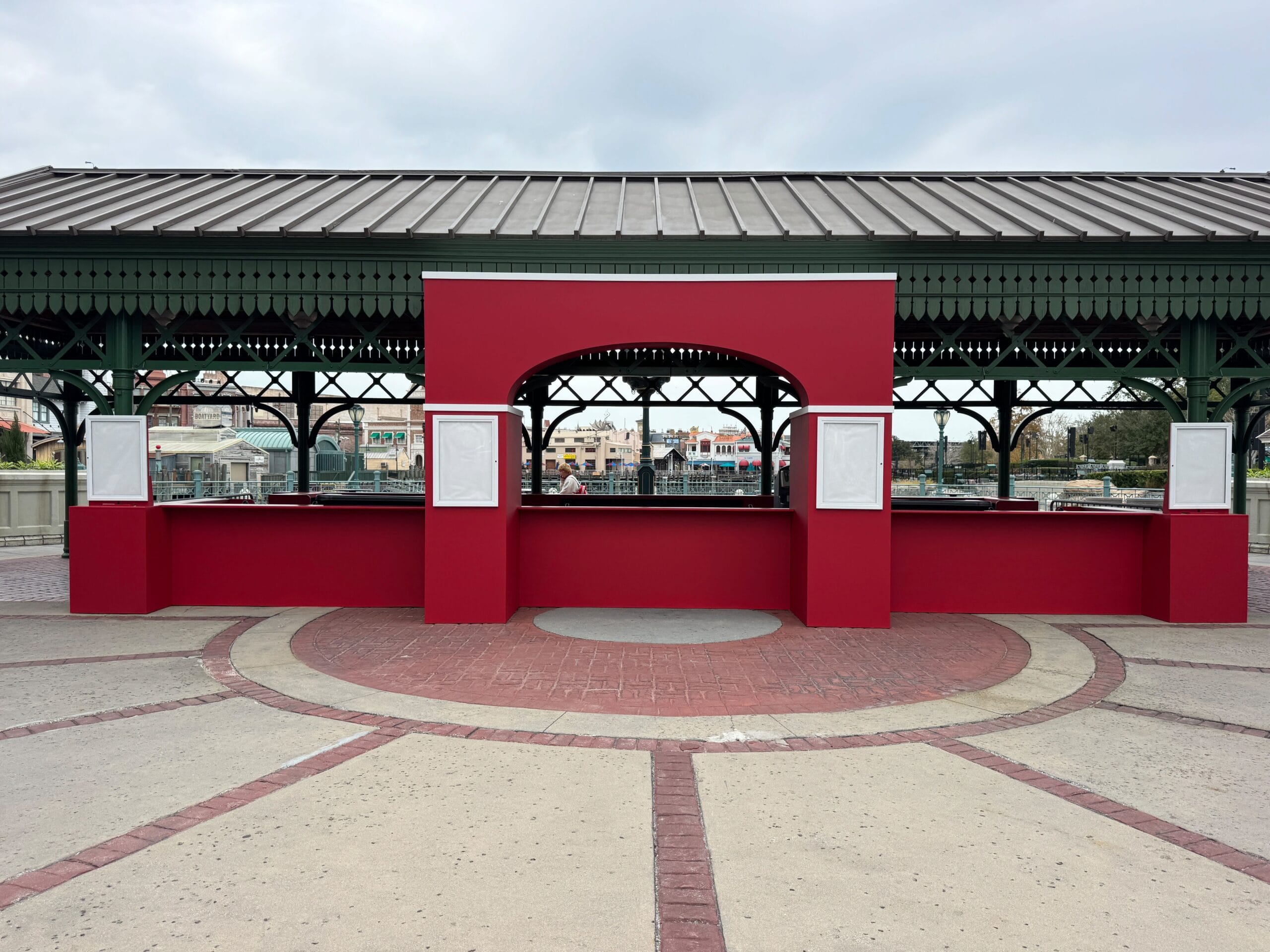 A red kiosk reminiscent of Mardi Gras flair stands under a green metal pavilion, its blank signboards waiting for a festive touch. A circular patterned concrete floor stretches out in front, hinting at vibrant food booths to come.