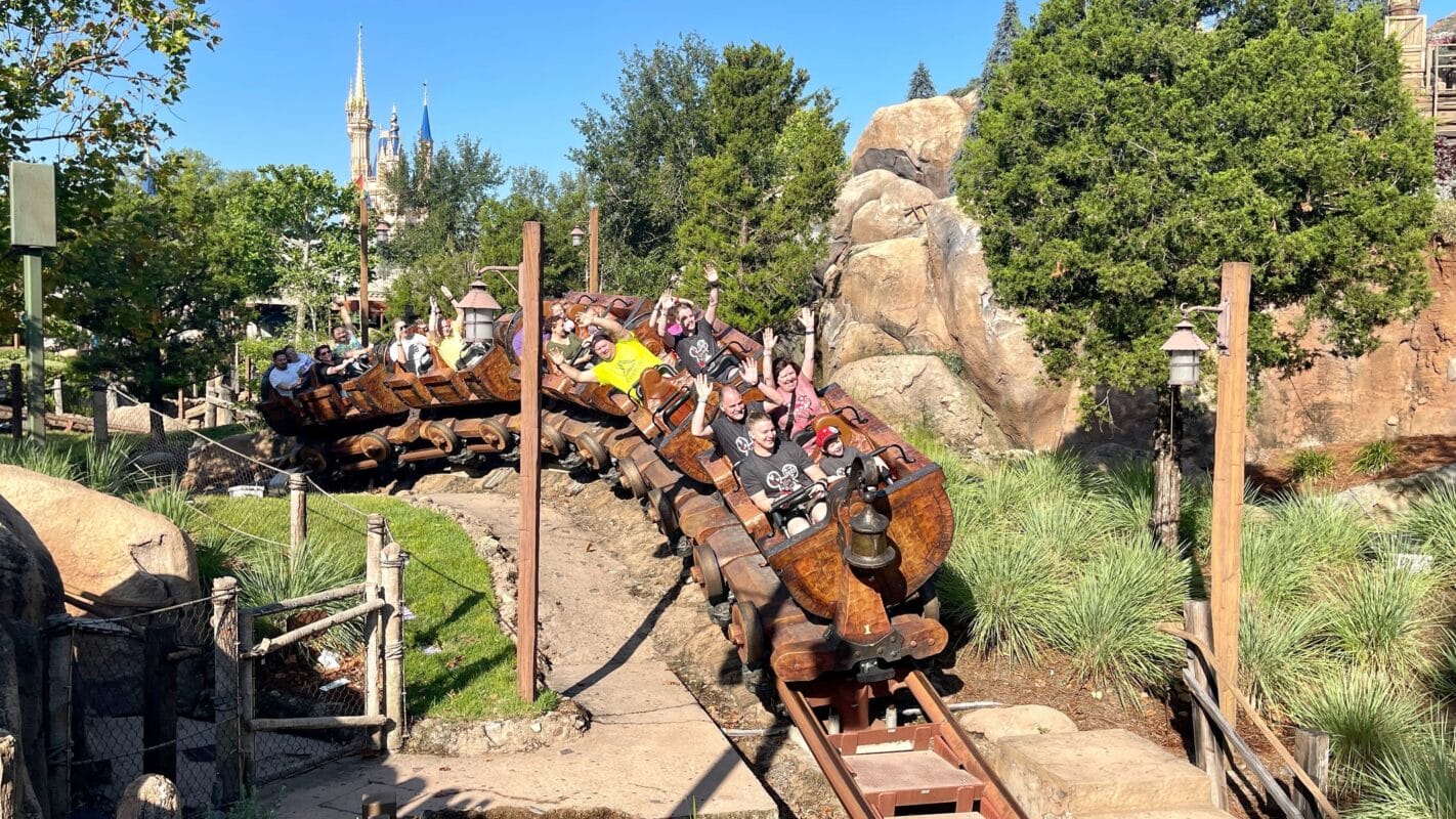 People enjoying the Seven Dwarfs Mine Train roller coaster, with trees and a castle in Magic Kingdom on a sunny day.