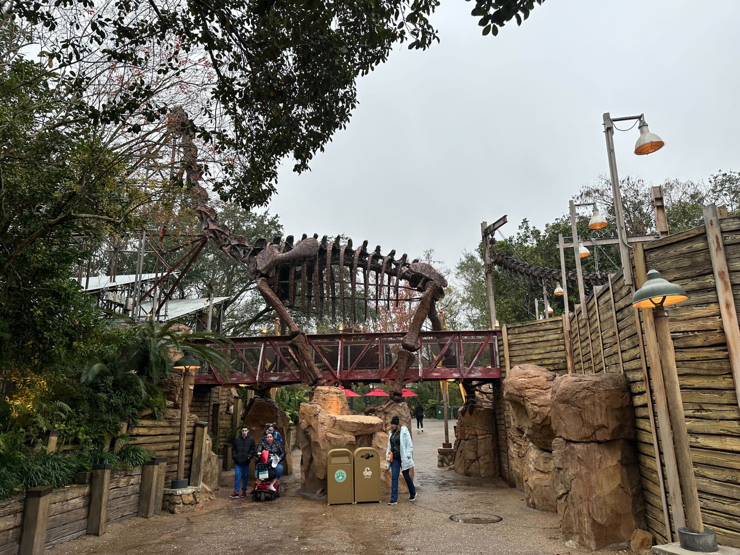 Entrance to a theme park with a large dinosaur skeleton structure above. A few people are standing at the entrance on a cloudy day.