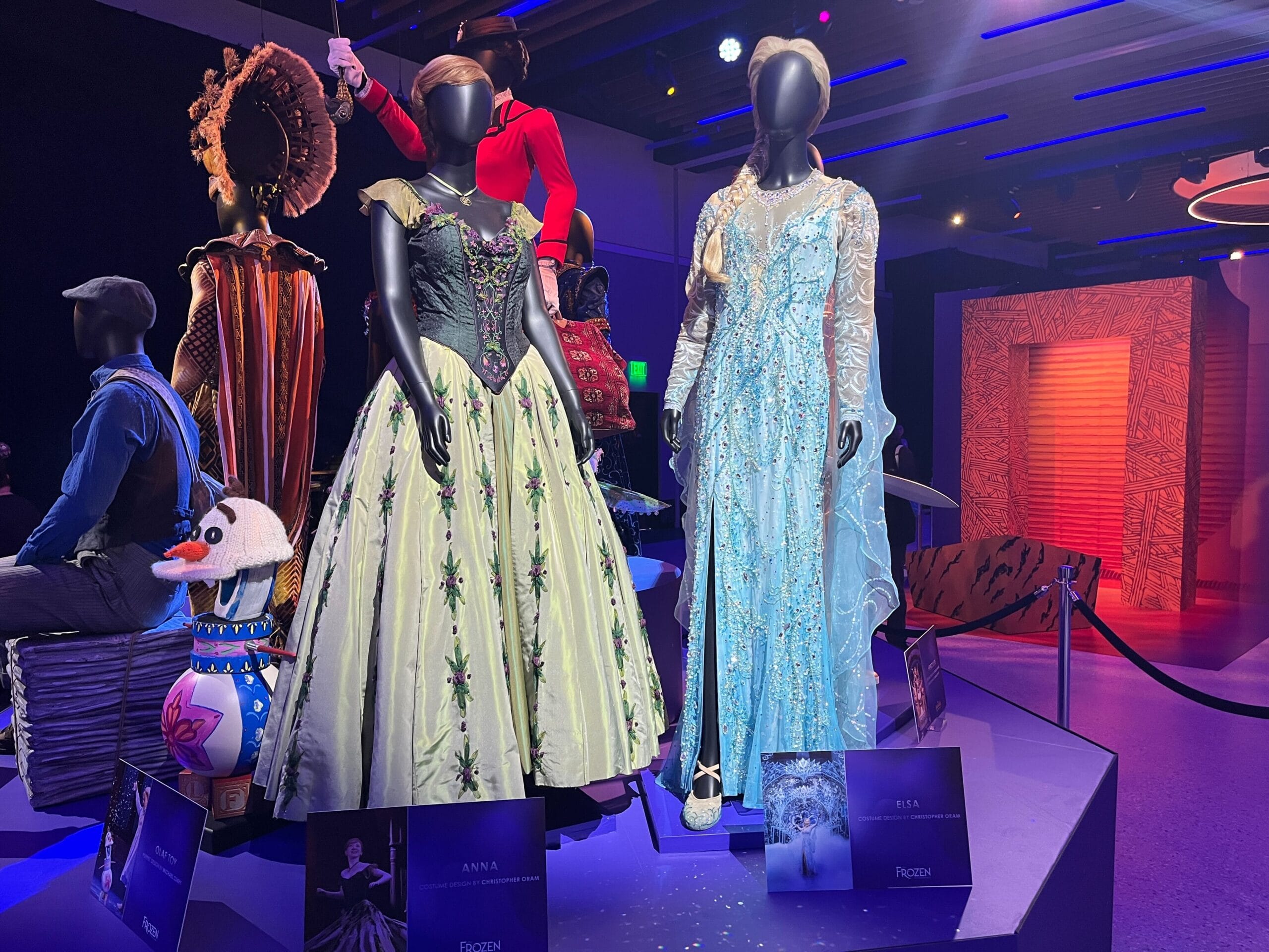 Mannequins display Anna and Elsa costumes from "Frozen" on a platform, surrounded by themed props and lighting in the enchanting Disney on Broadway Exhibit.