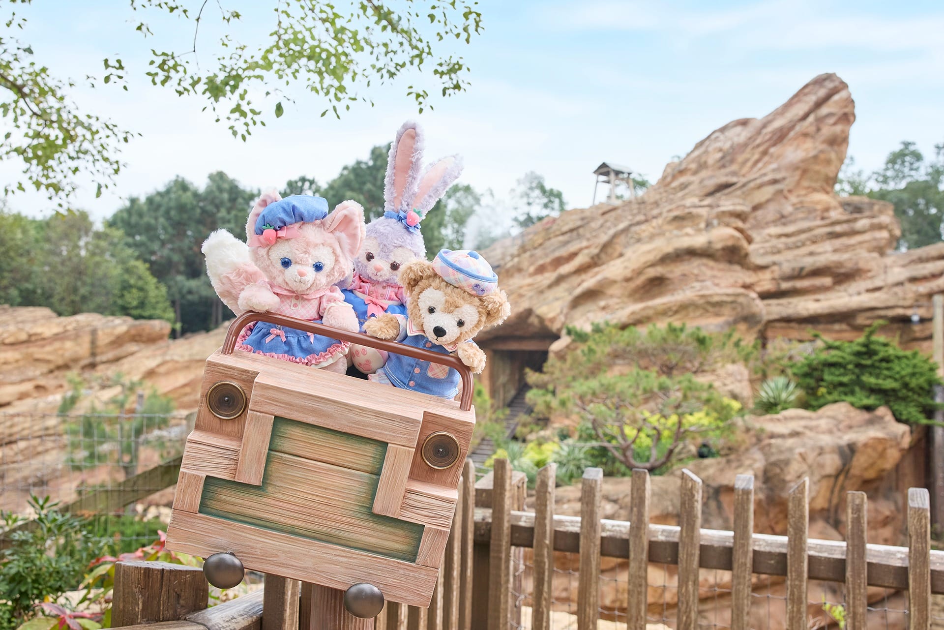 Three stuffed animals dressed in costumes ride in a wooden vehicle on a display, reminiscent of Play Days at Hong Kong Disneyland. A rocky landscape and trees form the enchanting background.