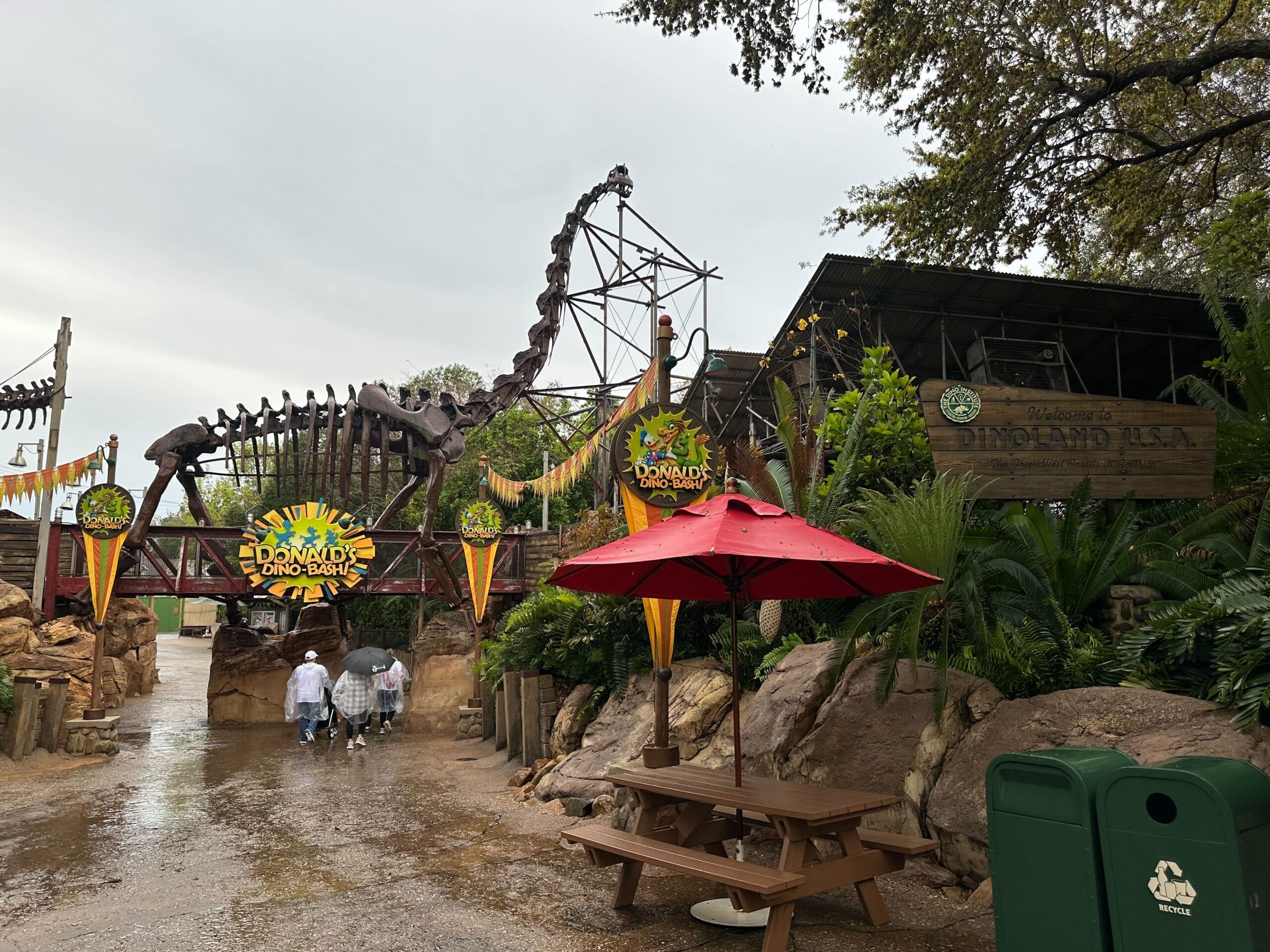 Entrance to a dinosaur-themed amusement area with skeletal decorations, vibrant signs, a red umbrella, picnic table, and trash cans, under overcast skies.