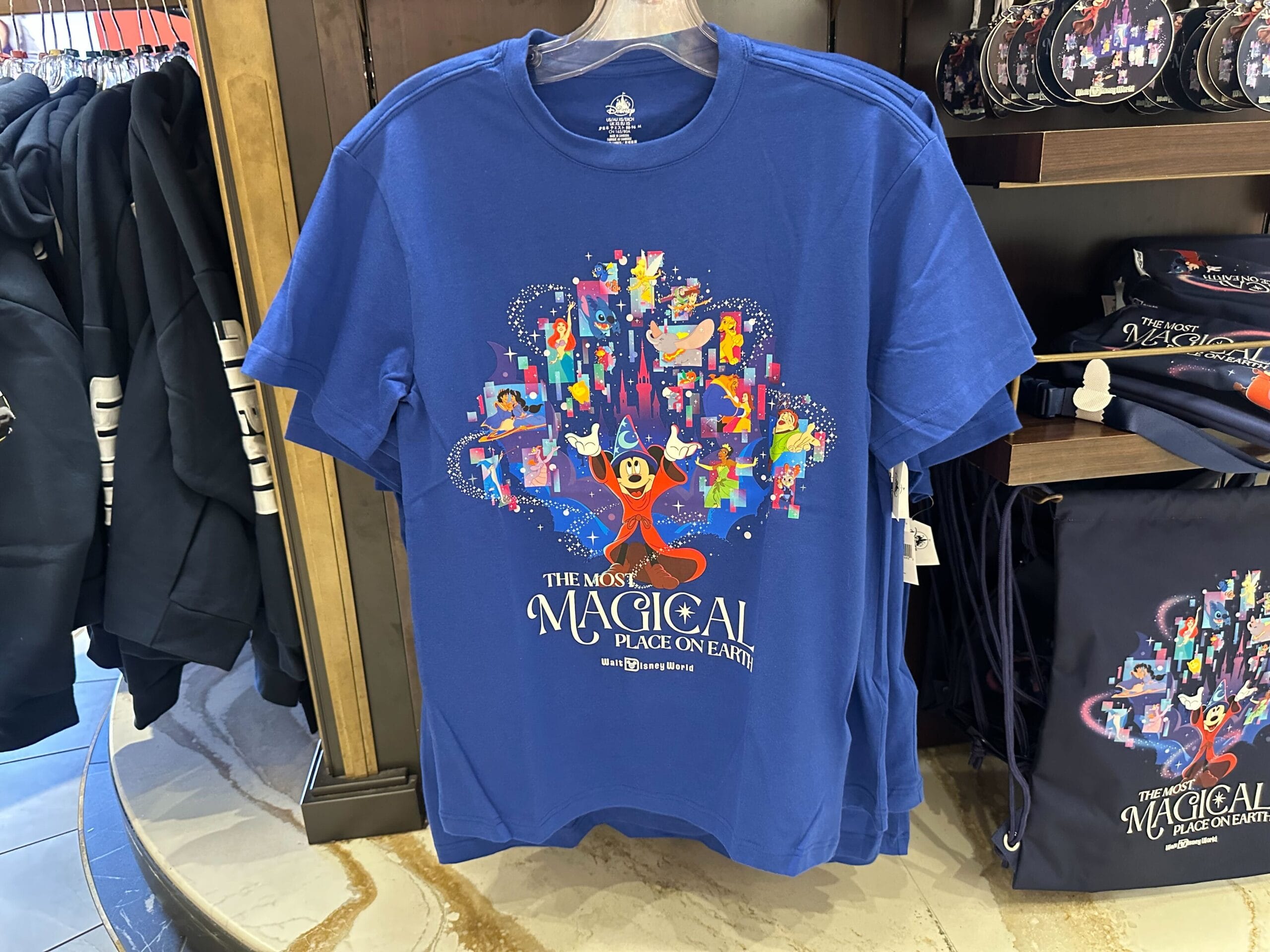 Blue T-shirt from the Apparel Collection showcases colorful illustrations of Disney characters, including Sorcerer Mickey, with the text "The Most Magical Place On Earth" in a store display.