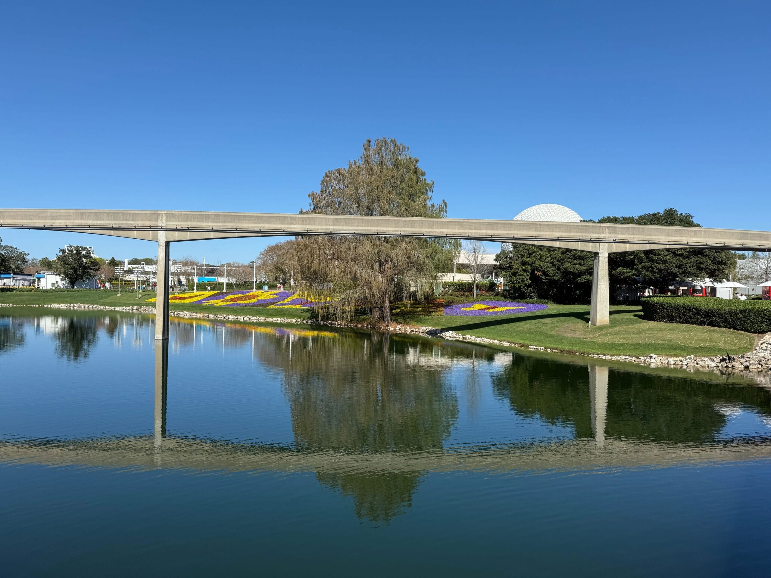 A monorail track glides over a reflective body of water, surrounded by vibrant flowerbeds inspired by the EPCOT Flower & Garden Festival, with trees in the background under a clear blue sky.