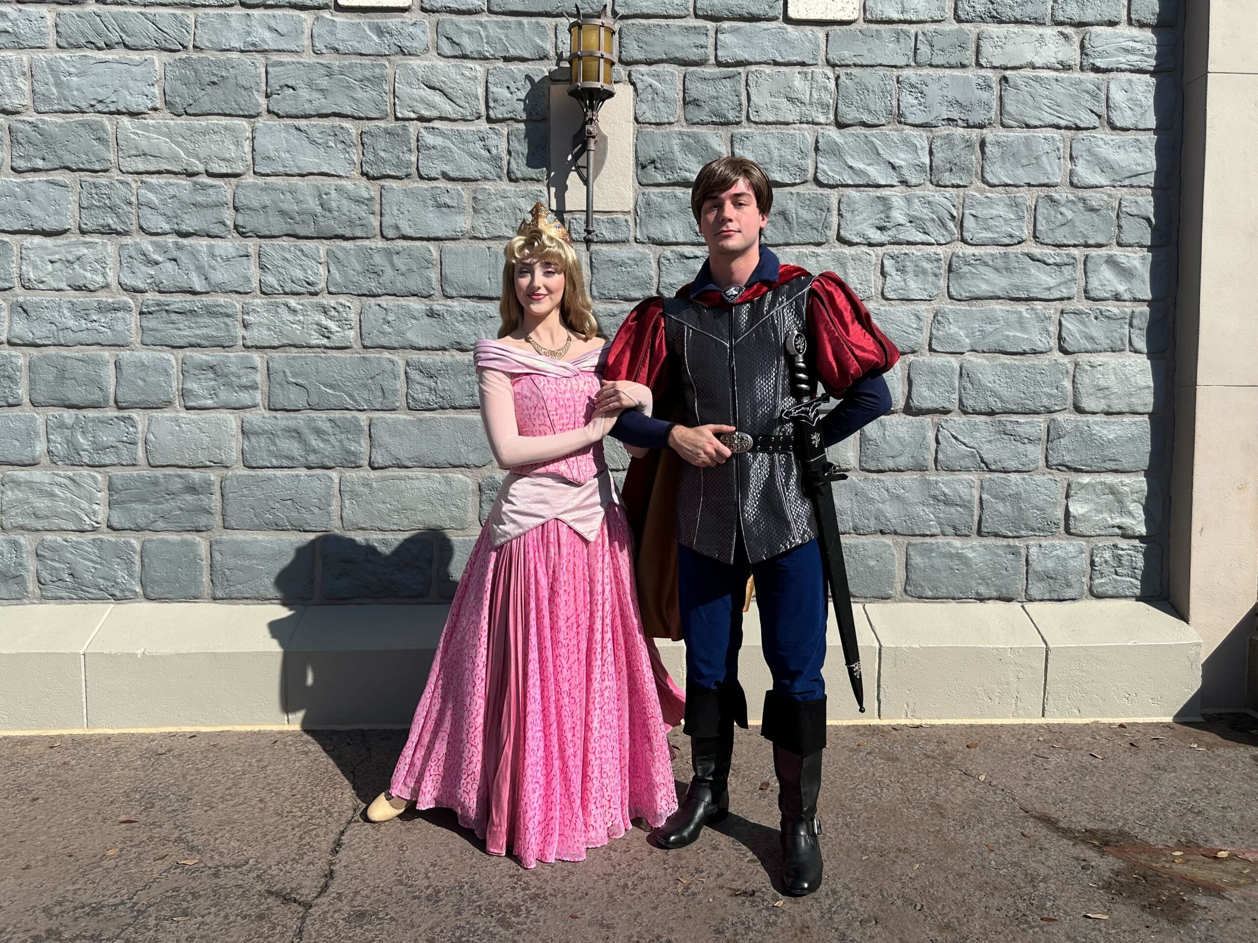 Two people dressed as a Disney couple strike a pose against a stone wall. One wears a pink dress and crown, while the other dons medieval armor with a red cape—capturing romance and magic just in time for Valentine's Day 2025.