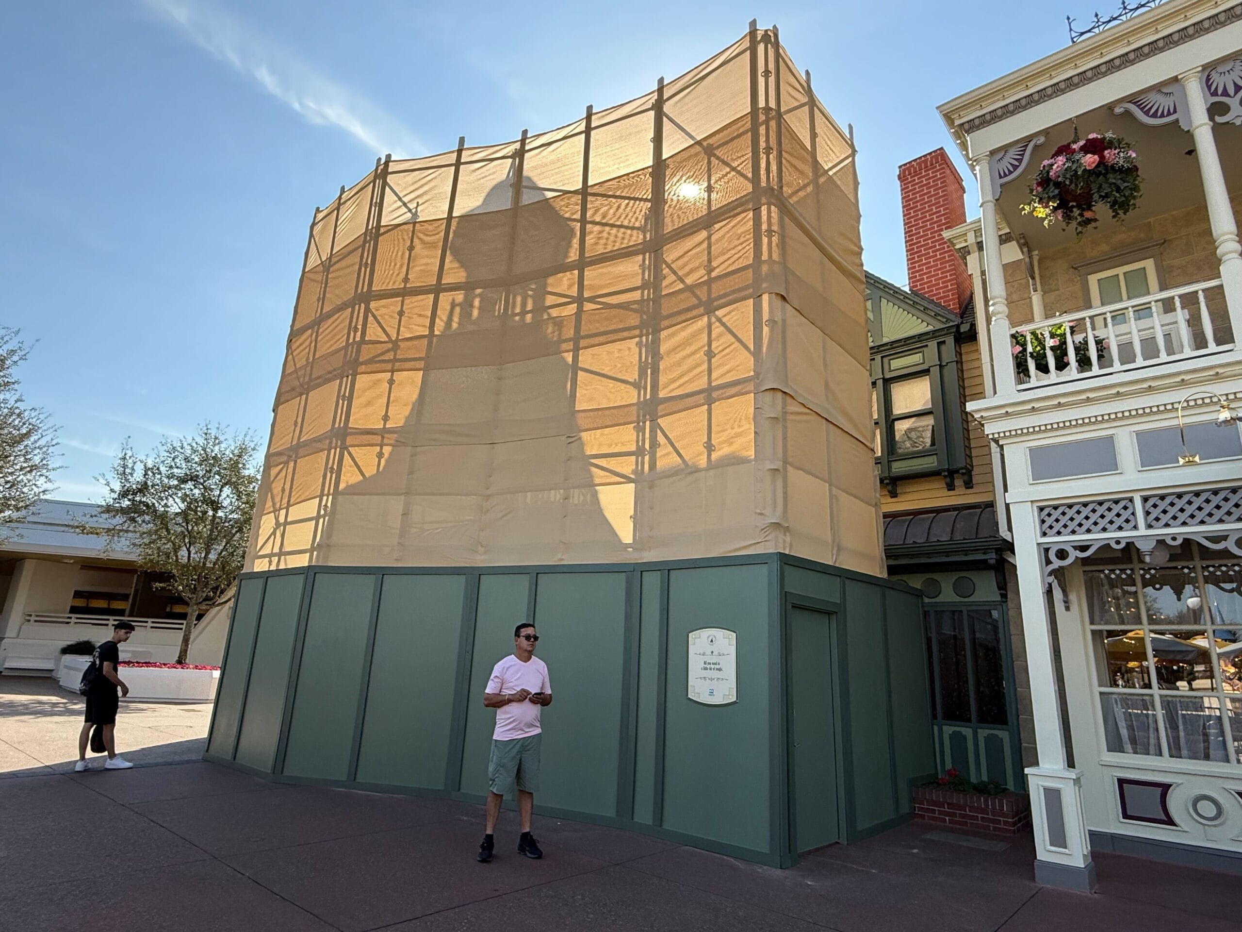 A man stands in front of a building covered with construction material and scaffolding, hinting at the upcoming transformation of the Plaza Restaurant. A sign is visible on the green barricade, adding intrigue to the scene.