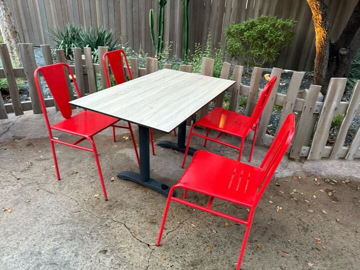 Four red chairs surround a wooden table on the concrete patio of Red’s Community Garden, enclosed by a wooden fence with lush plants in the background.
