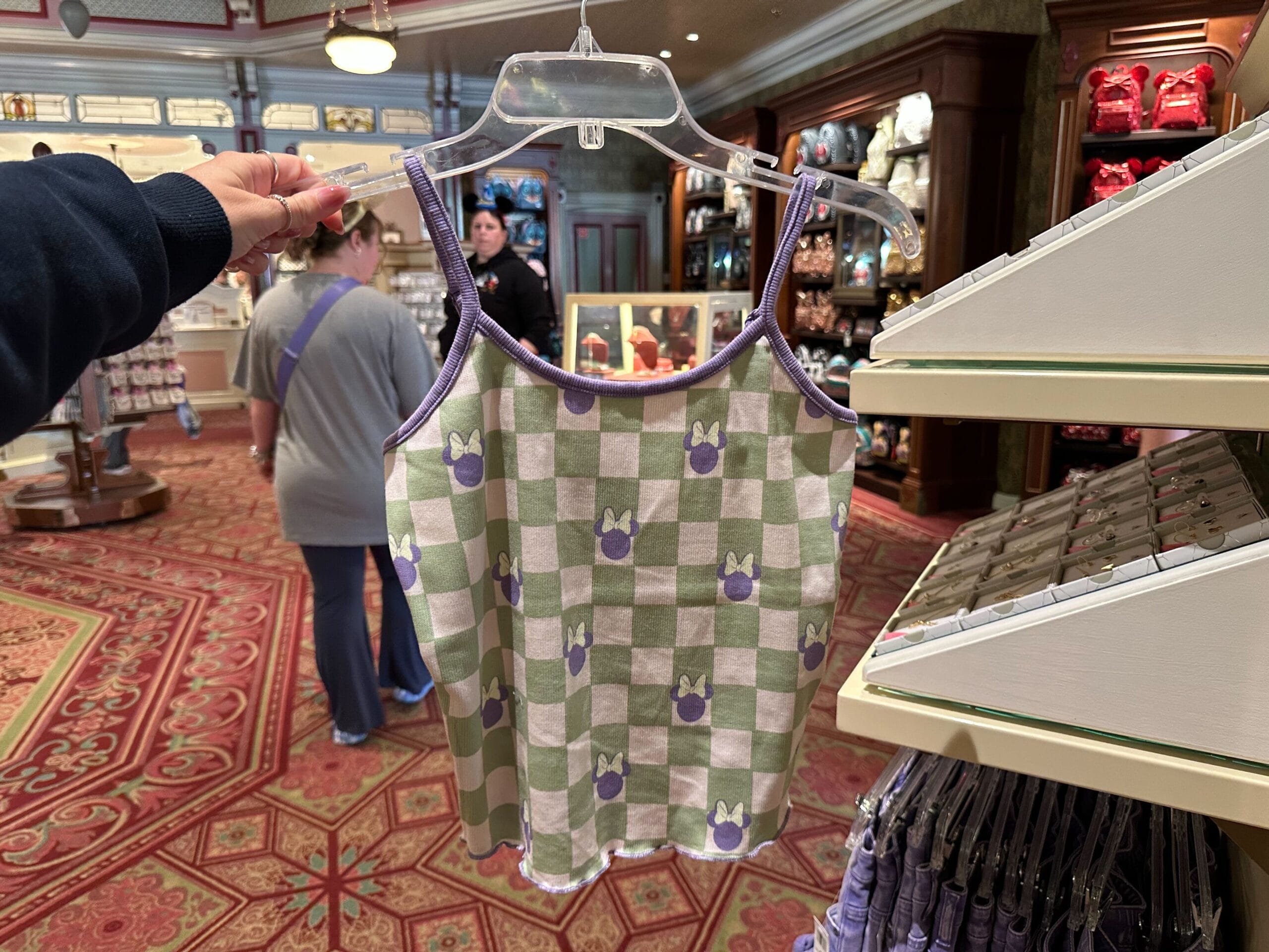 A green and white checkered tank top with purple trim and bow designs, part of the Minnie Mouse Collection, is held on a hanger in a store. Shelves with merchandise and two people are seen in the background.