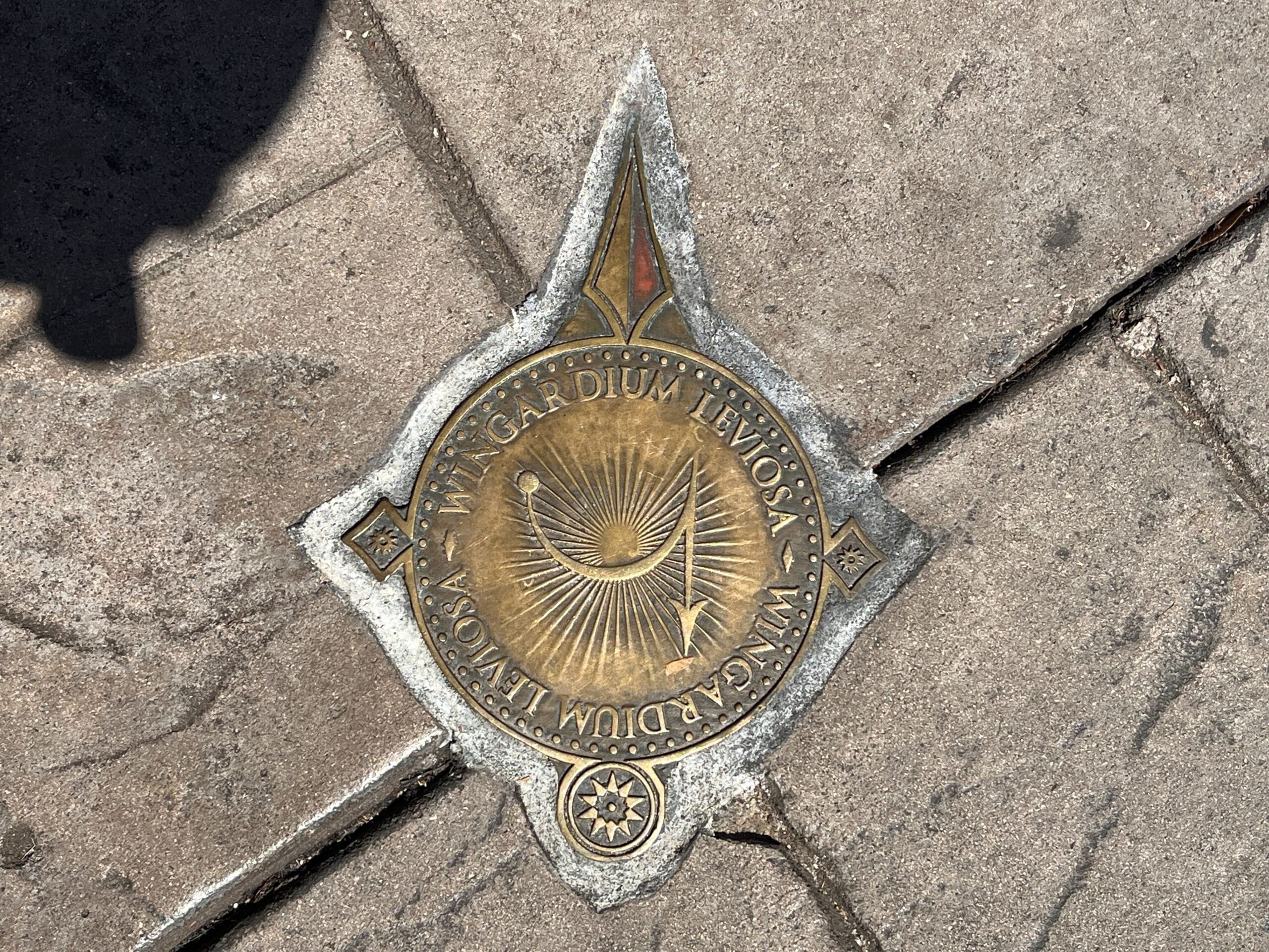 A bronze plaque, part of the spell locations series, is embedded in the pavement, featuring the text "Wingardium Leviosa" alongside a delicate feather design.