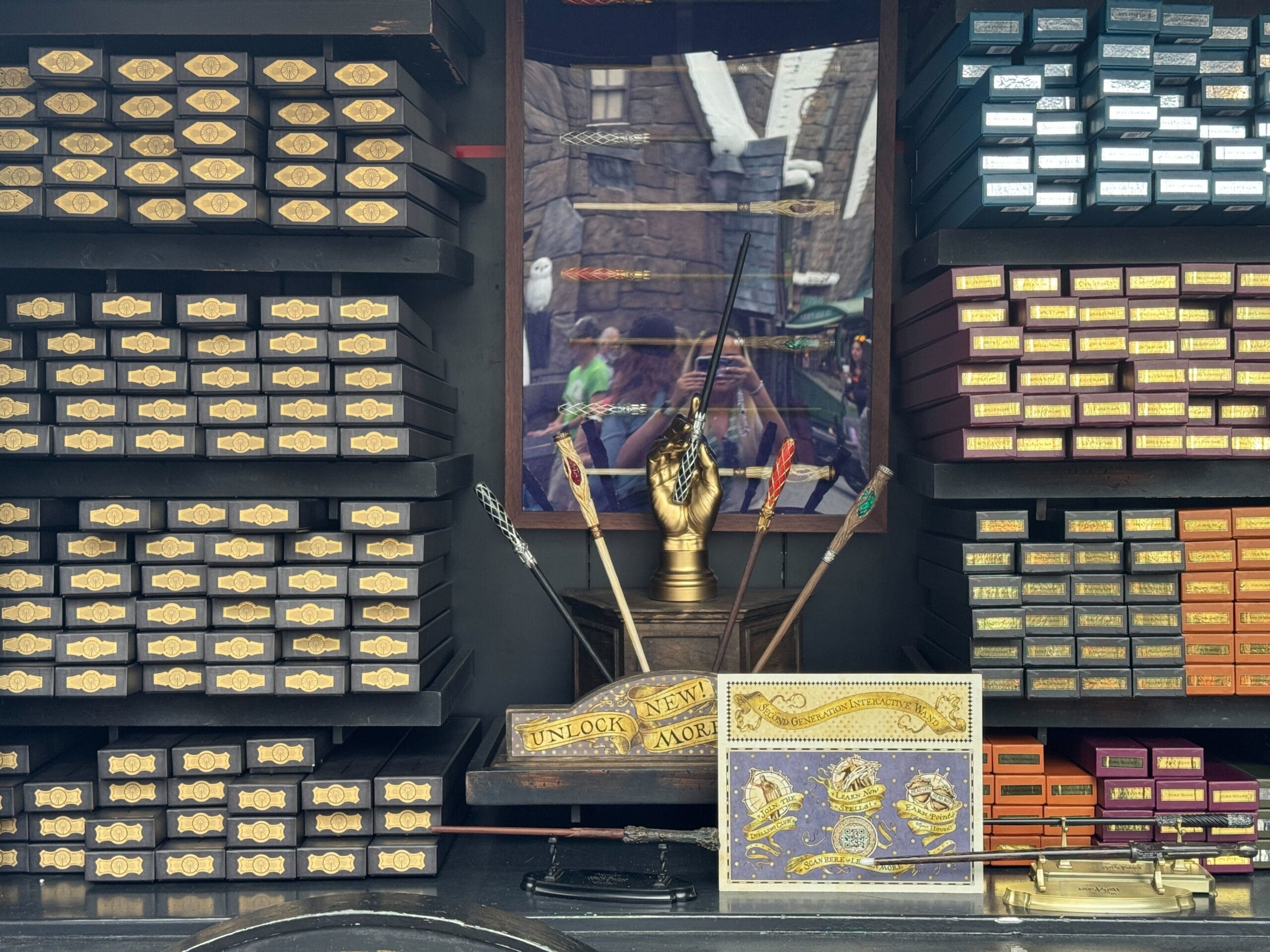 A shop display features neatly stacked boxes on shelves, a central exhibit of quill pens in a golden hand, and decorative items. Among them is a sign that reads "Unlock New Magic," inviting you to discover the allure of Interactive Wands.