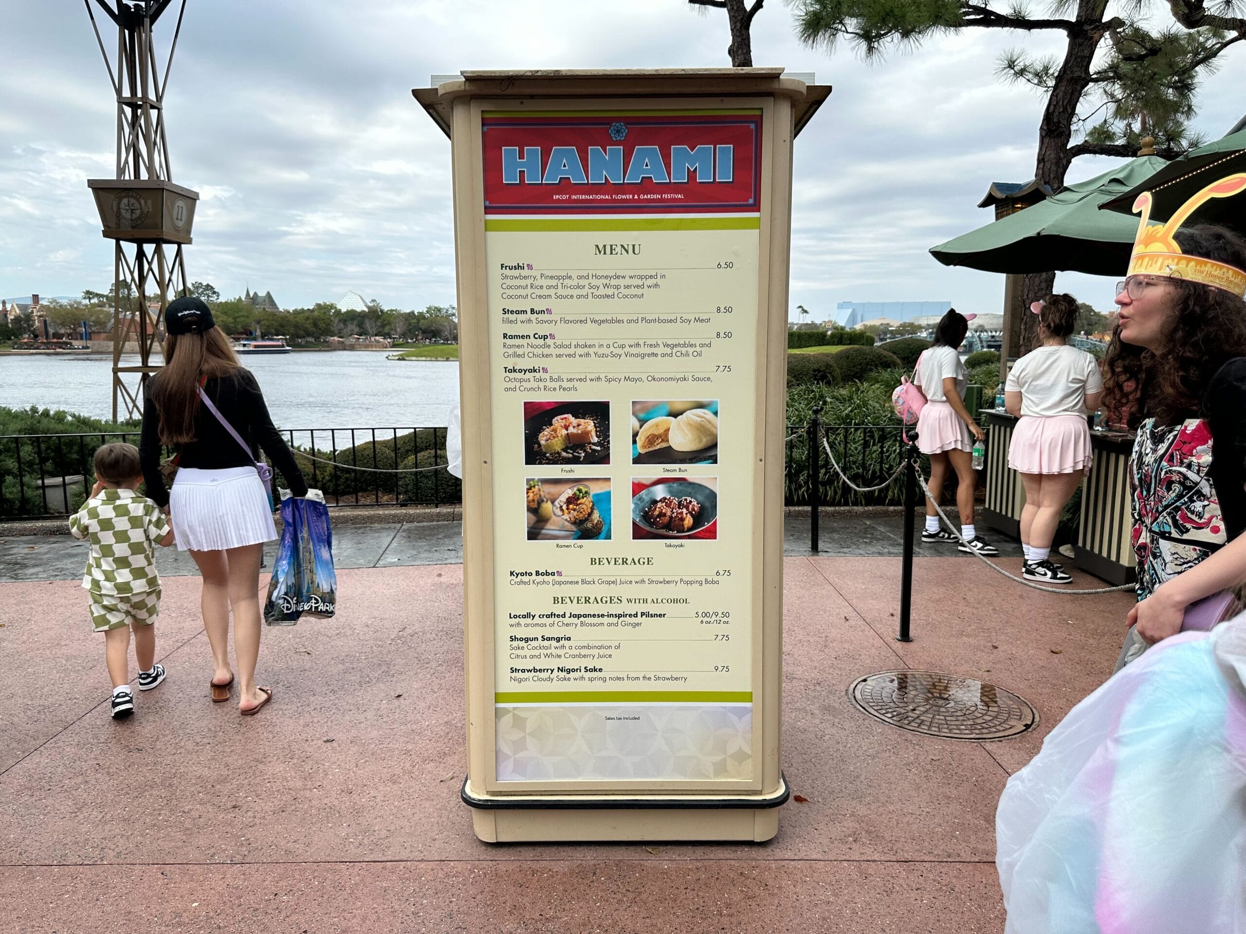 A menu board titled "Hanami" showcases various food and drink options, capturing the essence of a Flower & Garden Festival. In this vibrant outdoor scene, people stroll by, including a child on the left and someone donning a festive crown on the right.