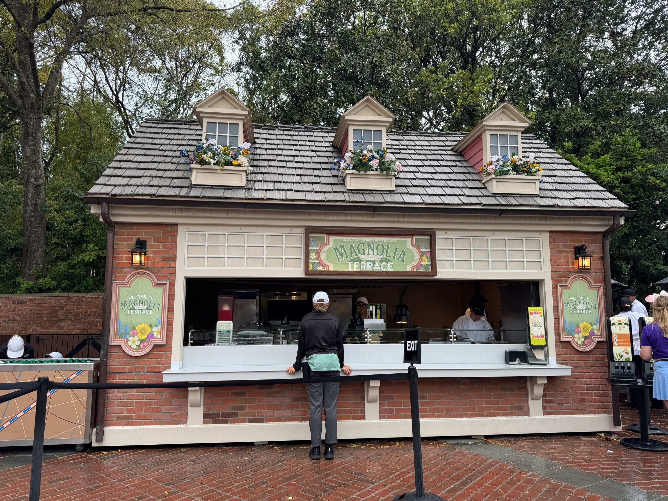 A person stands at the counter of Magnolia Terrace, a brick food kiosk adorned with three dormer windows and vibrant flower boxes, perfectly blending with the lush surroundings during the Flower & Garden Festival.